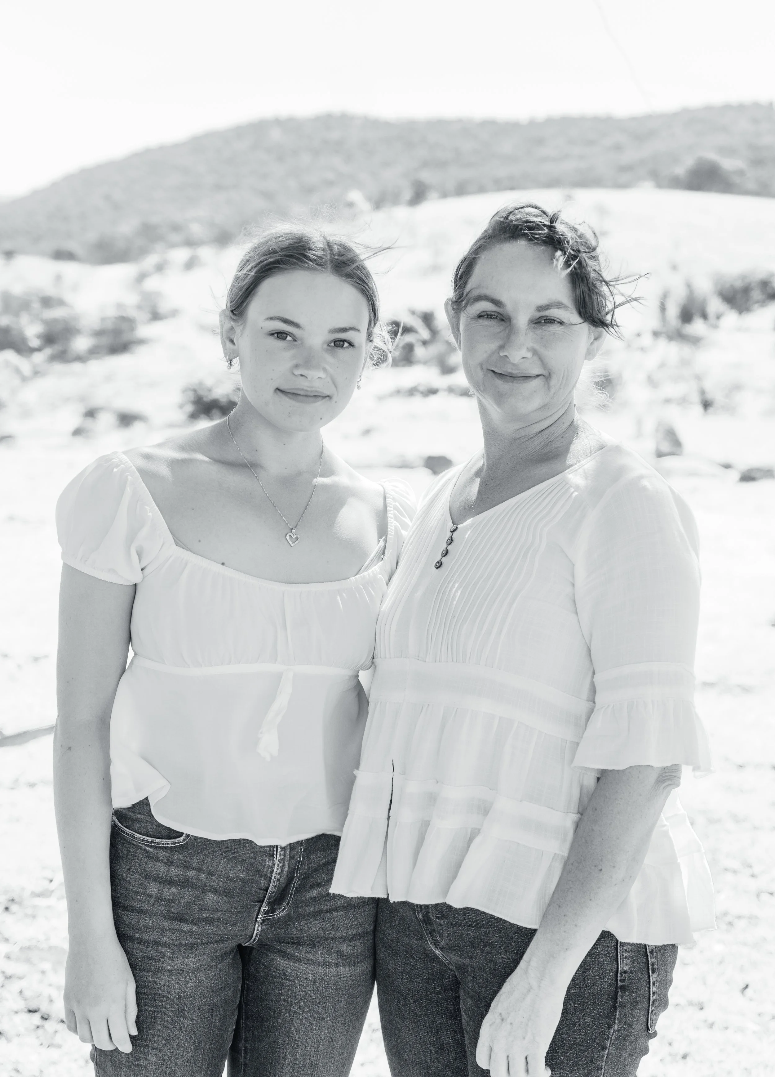 Two women standing outdoors in a sunny, mountainous landscape, wearing light-colored tops and jeans, smiling at the camera.