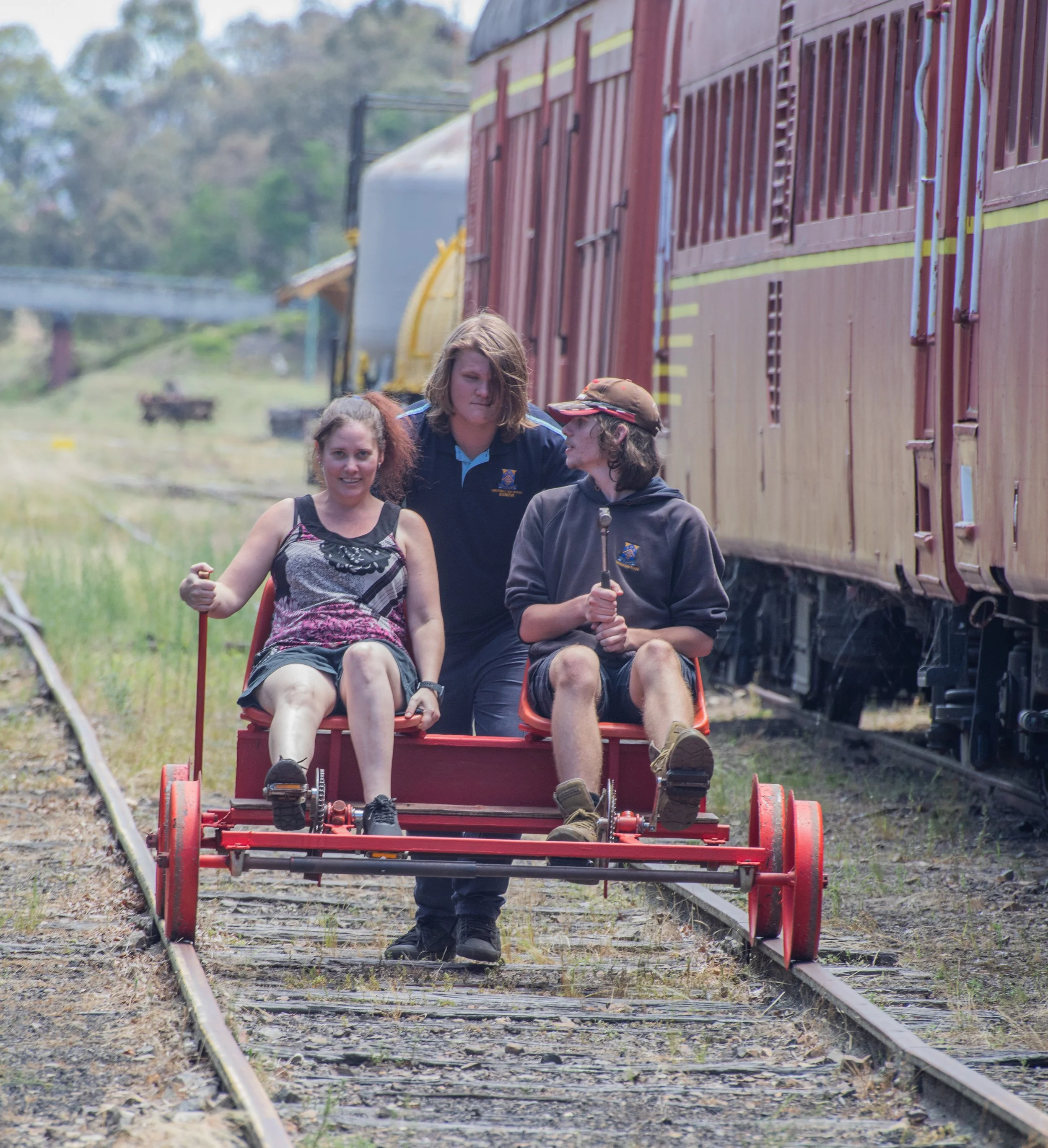 Three young people, two sitting and one standing, on a small rail cart beside train cars outdoors. The person on the left is a girl with curly hair wearing a sleeveless top, the girl in the middle is standing with blonde hair in a blue polo shirt, an