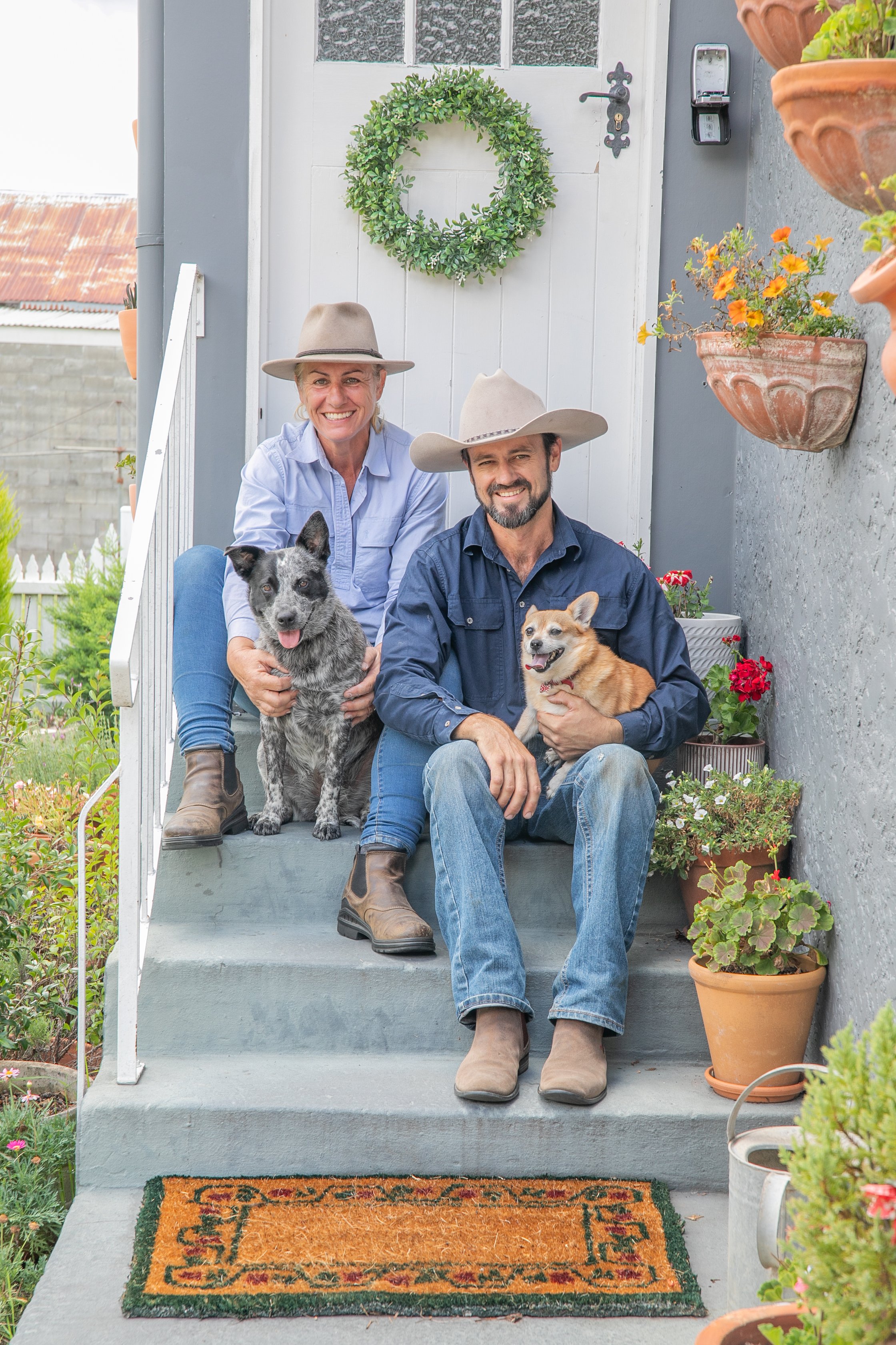 A happy older couple sitting on their front porch steps with two dogs, surrounded by potted plants and garden flowers, in front of a white door with a green wreath.