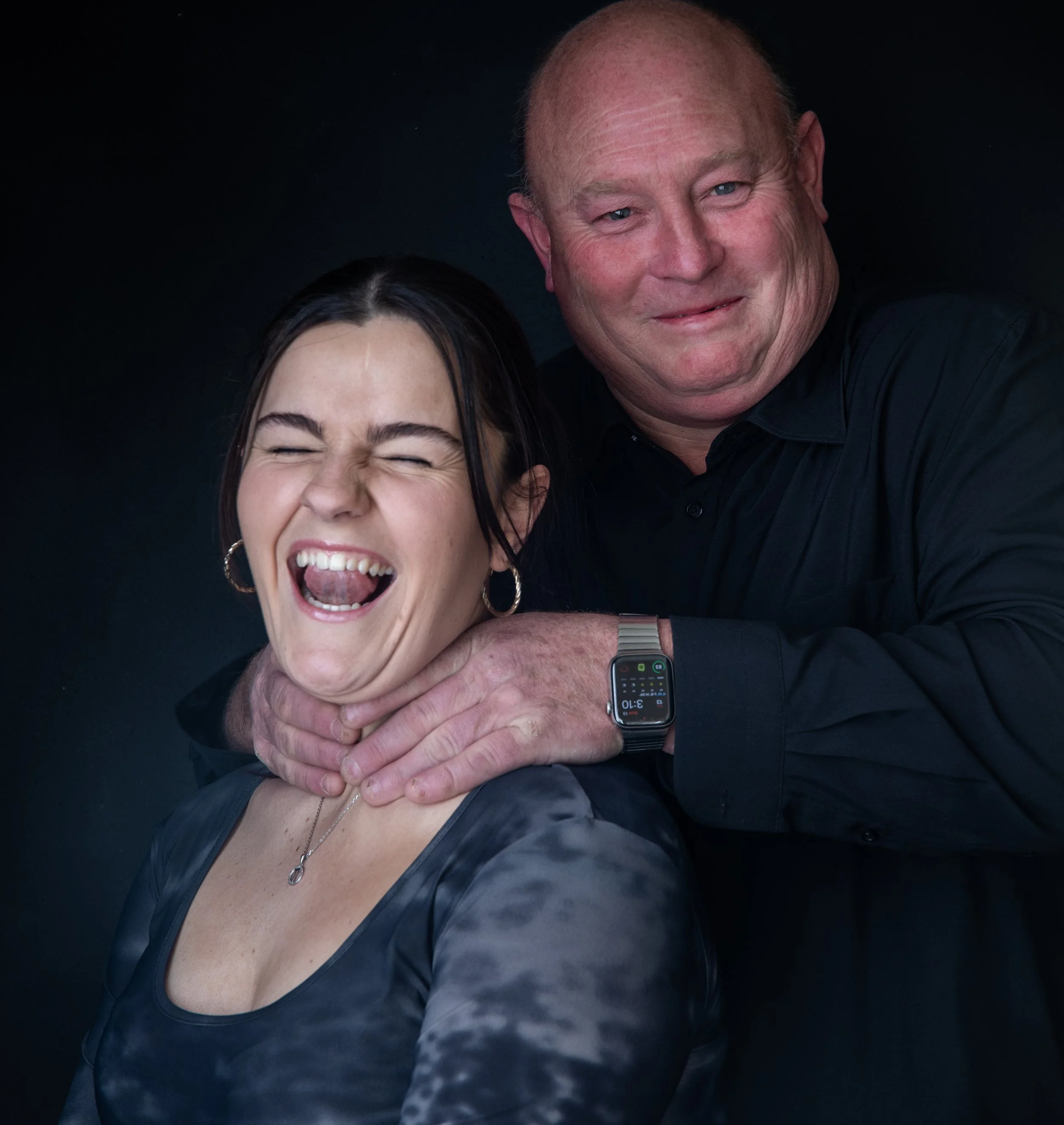 A man with a smile wearing a black shirt and a smartwatch on his wrist is holding a woman's neck from behind, and both are smiling and laughing, against a dark background.