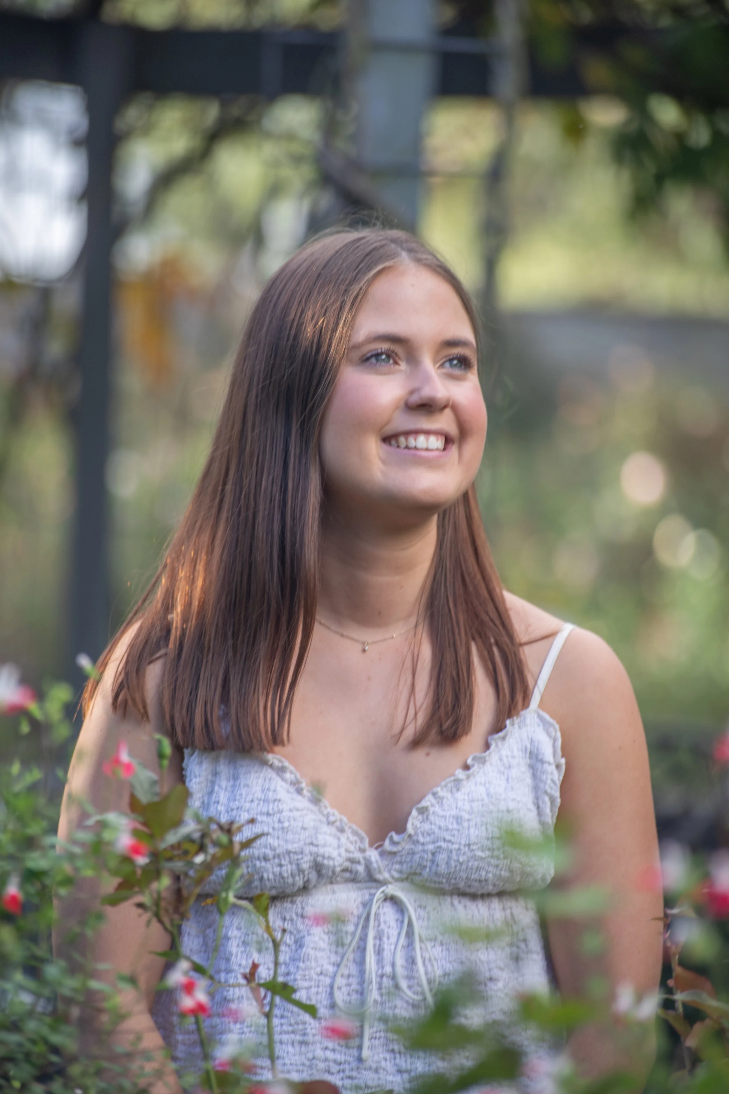 A young woman with brown hair smiling outdoors among greenery and pink flowers.