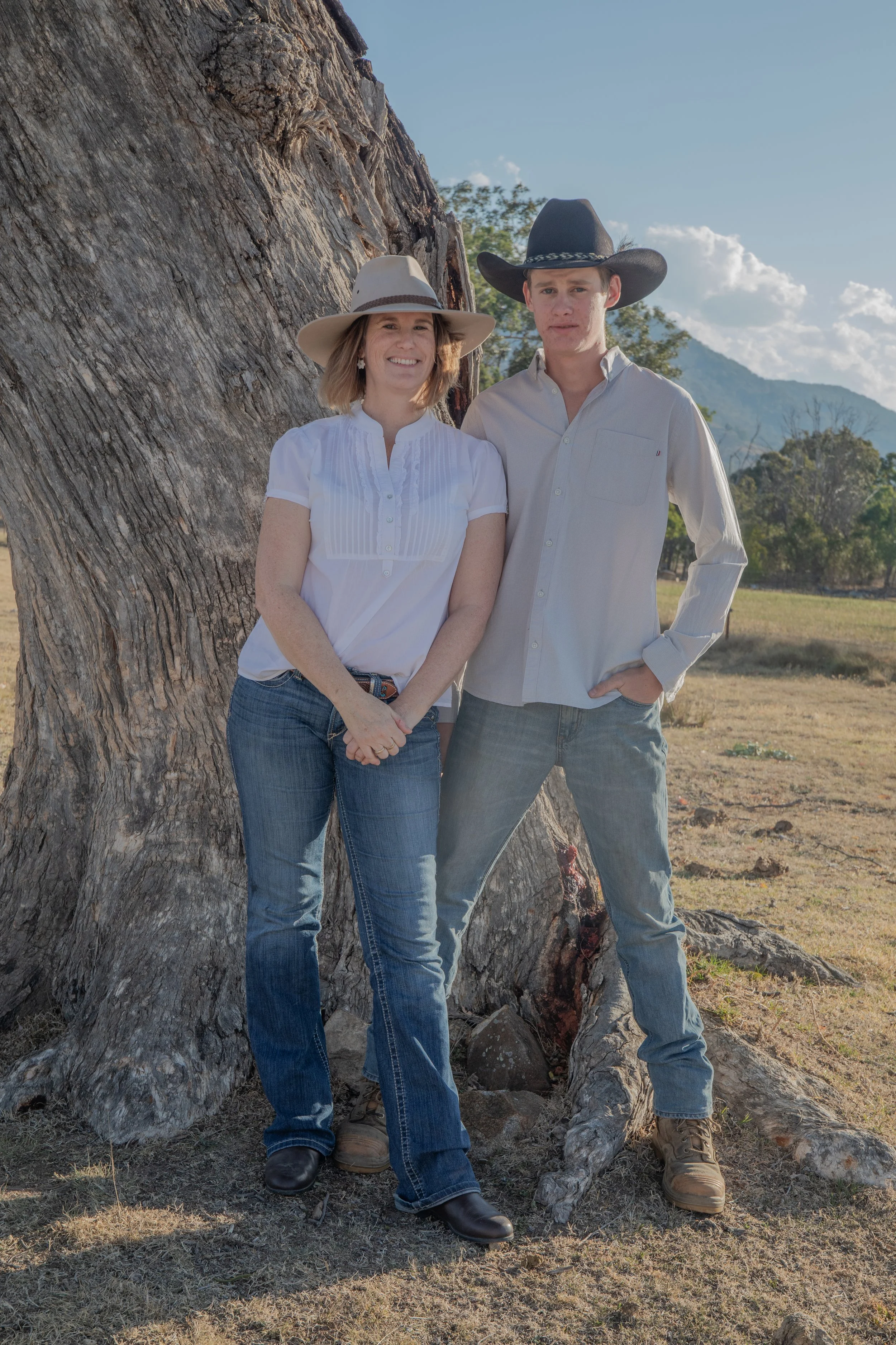 A woman and a man standing outdoors next to a large tree with mountains in the background, both wearing cowboy hats and casual clothing.