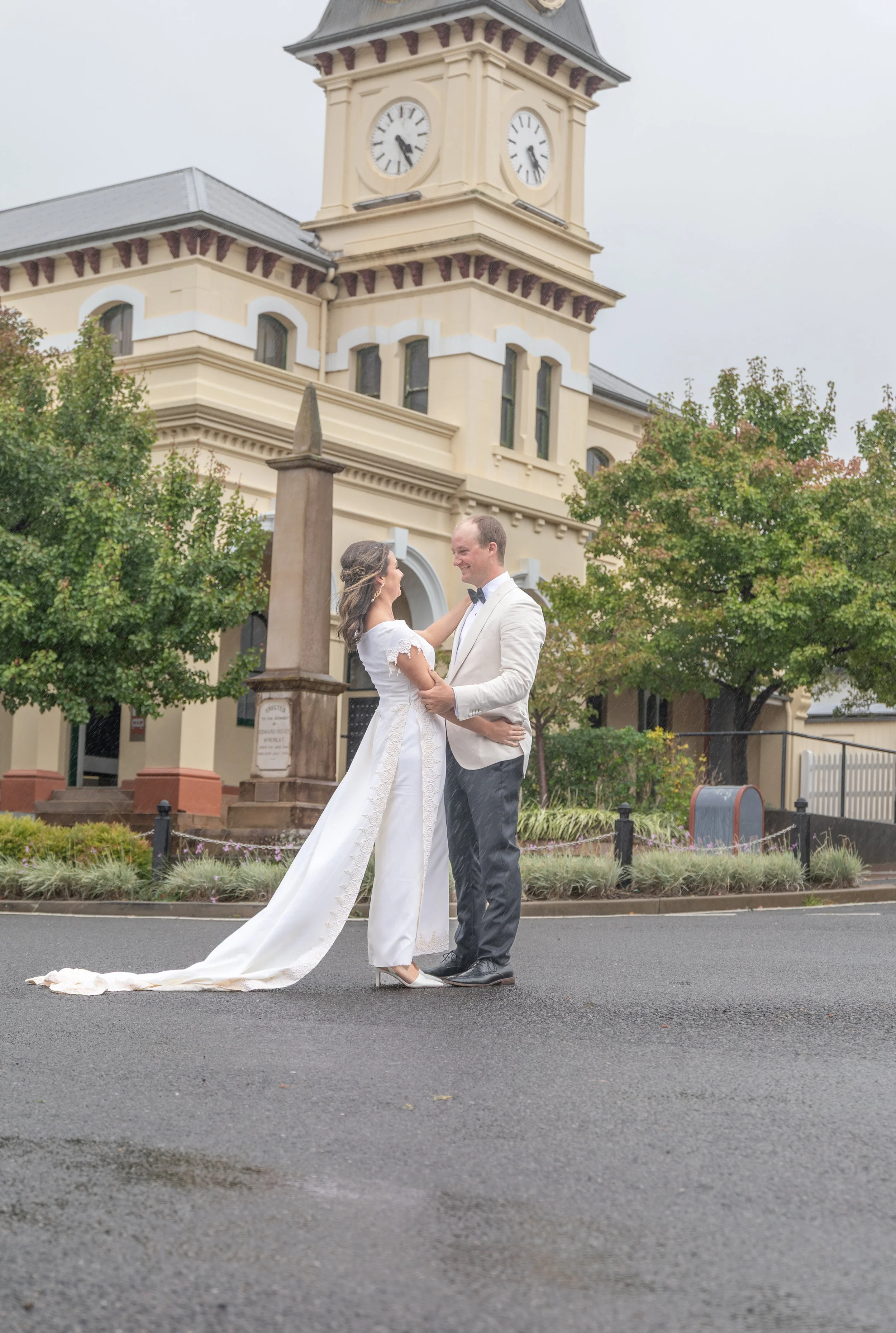 A newlywed couple dancing in the street in front of a historic building with a clock tower, trees, and greenery, on a cloudy day.