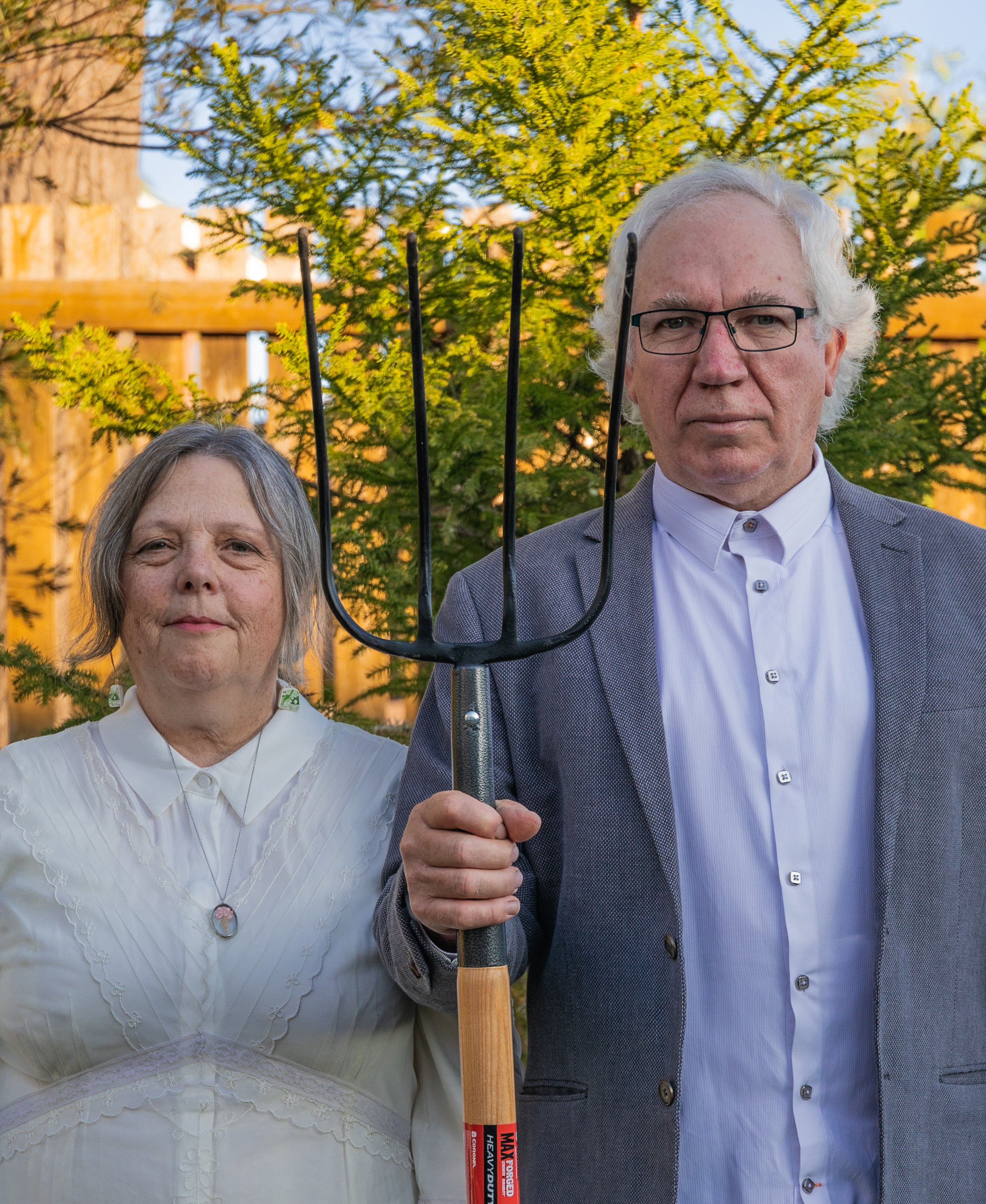 An elderly man and woman standing outdoors in front of a wooden fence and greenery, with the man holding a garden rake.