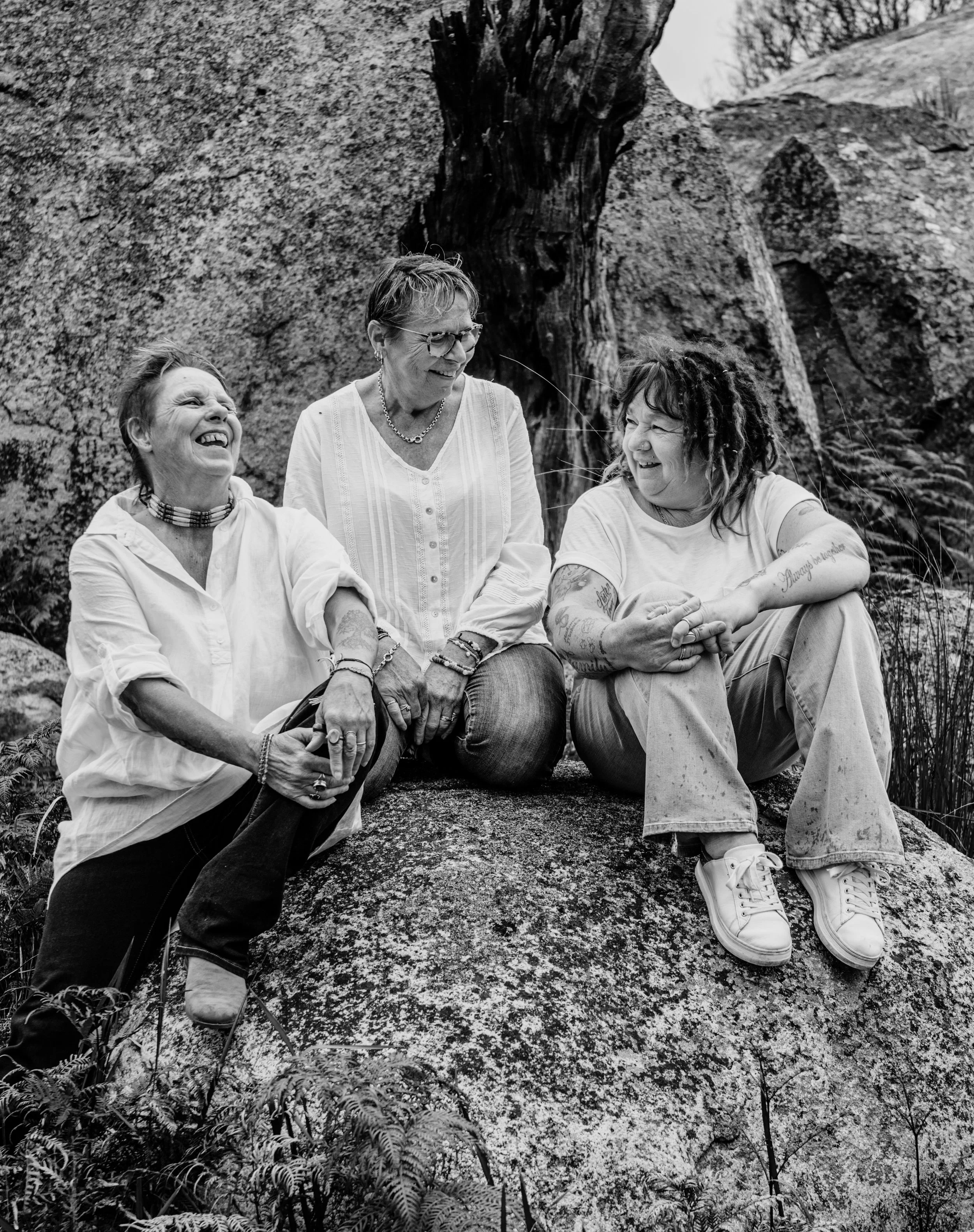 Three women sit on a large rock outdoors, laughing and smiling.