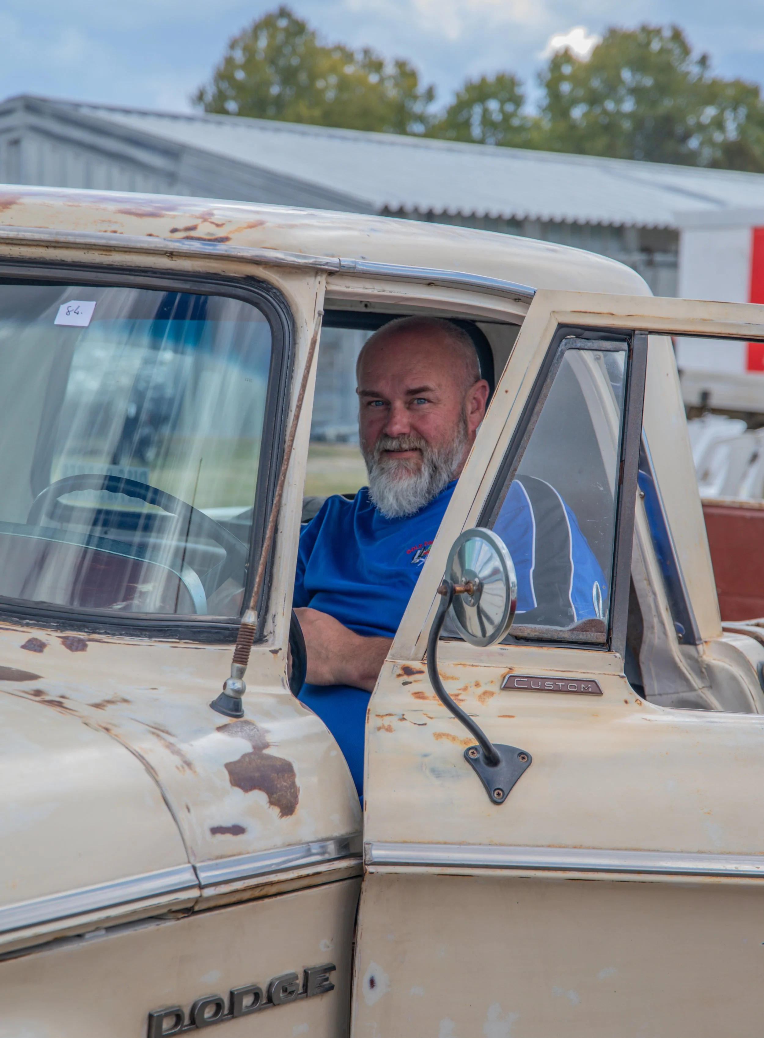 A man with a beard and blue shirt sitting in the driver's seat of an old, weathered Dodge pickup truck.