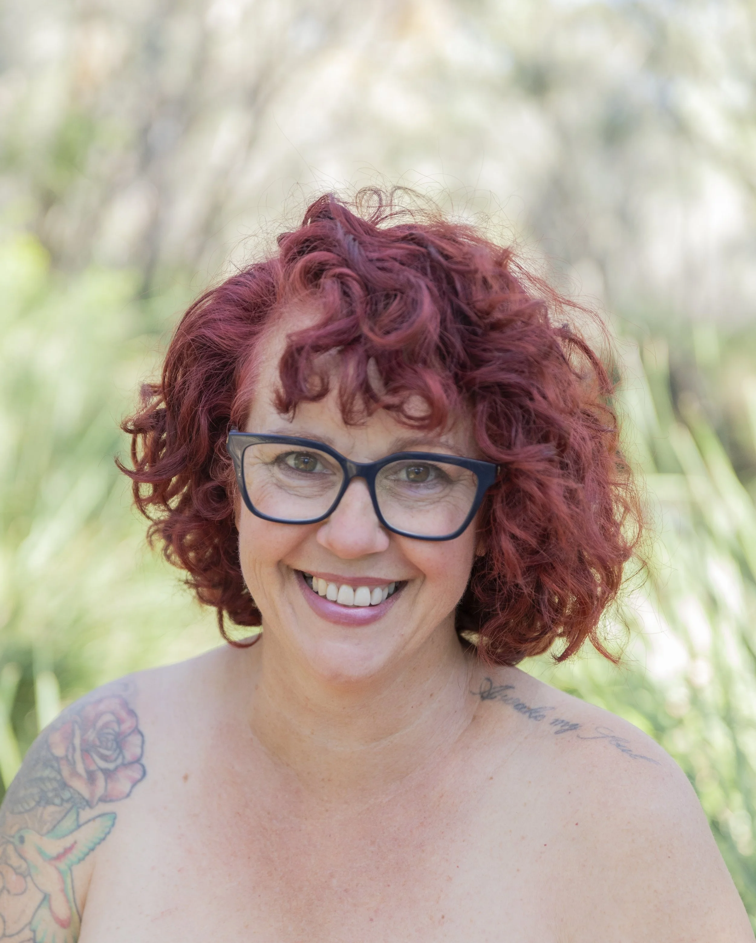 A smiling woman with curly red hair, wearing glasses, outdoors with greenery in the background.