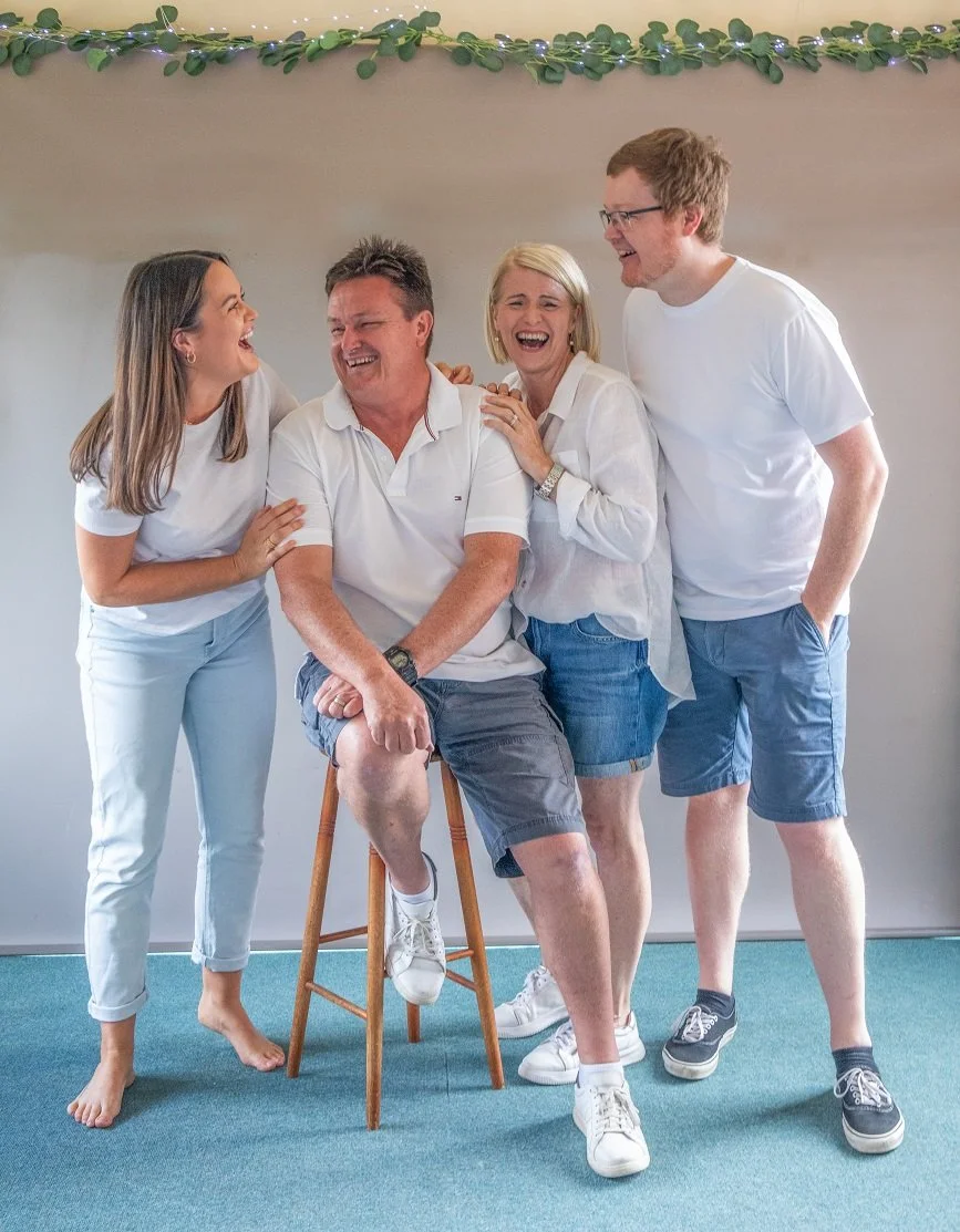 Four adults laughing and smiling together, with one seated on a wooden stool, in a room decorated with greenery and string lights.