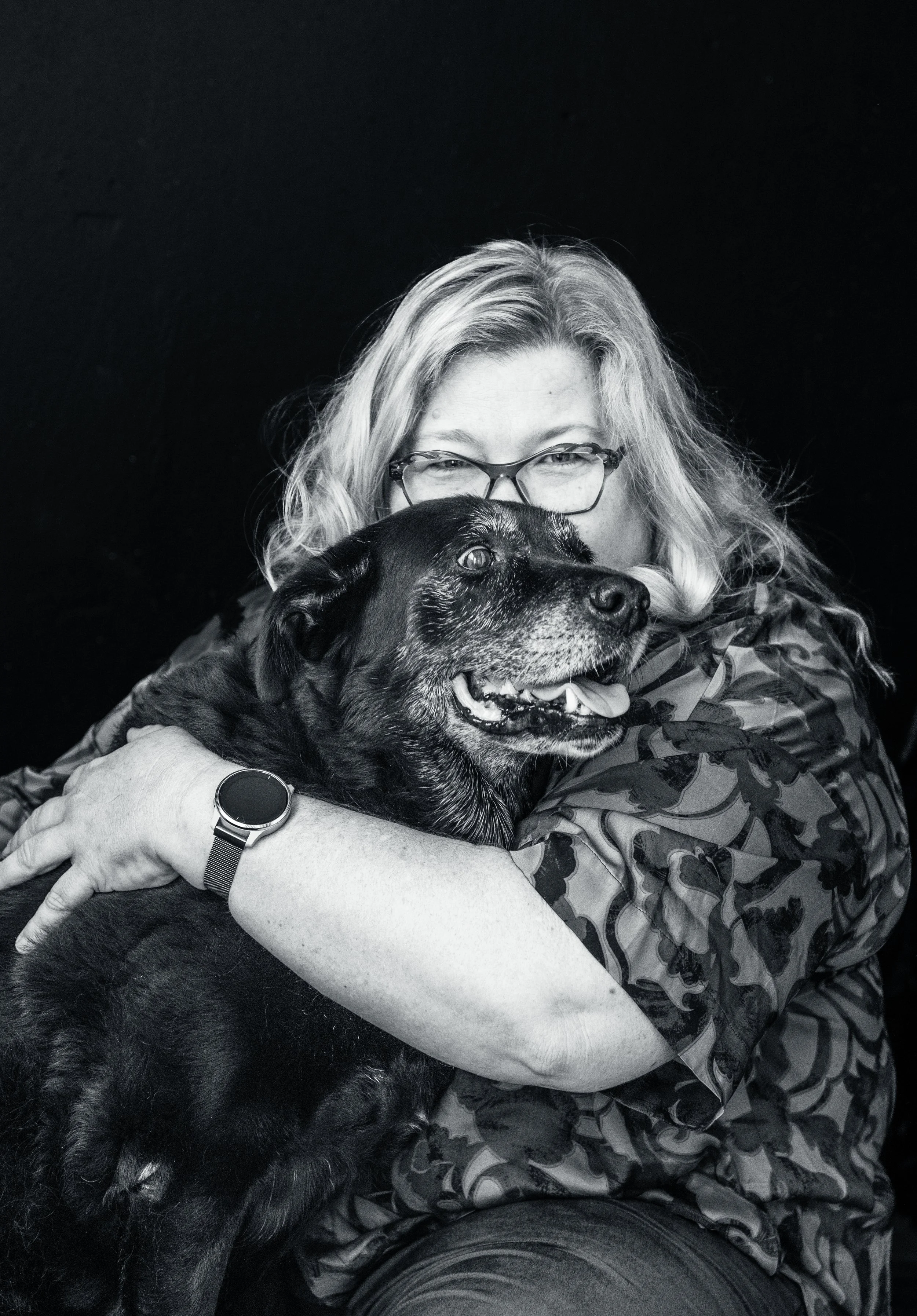 A woman with glasses hugging a black dog with gray fur on its face, against a dark background.
