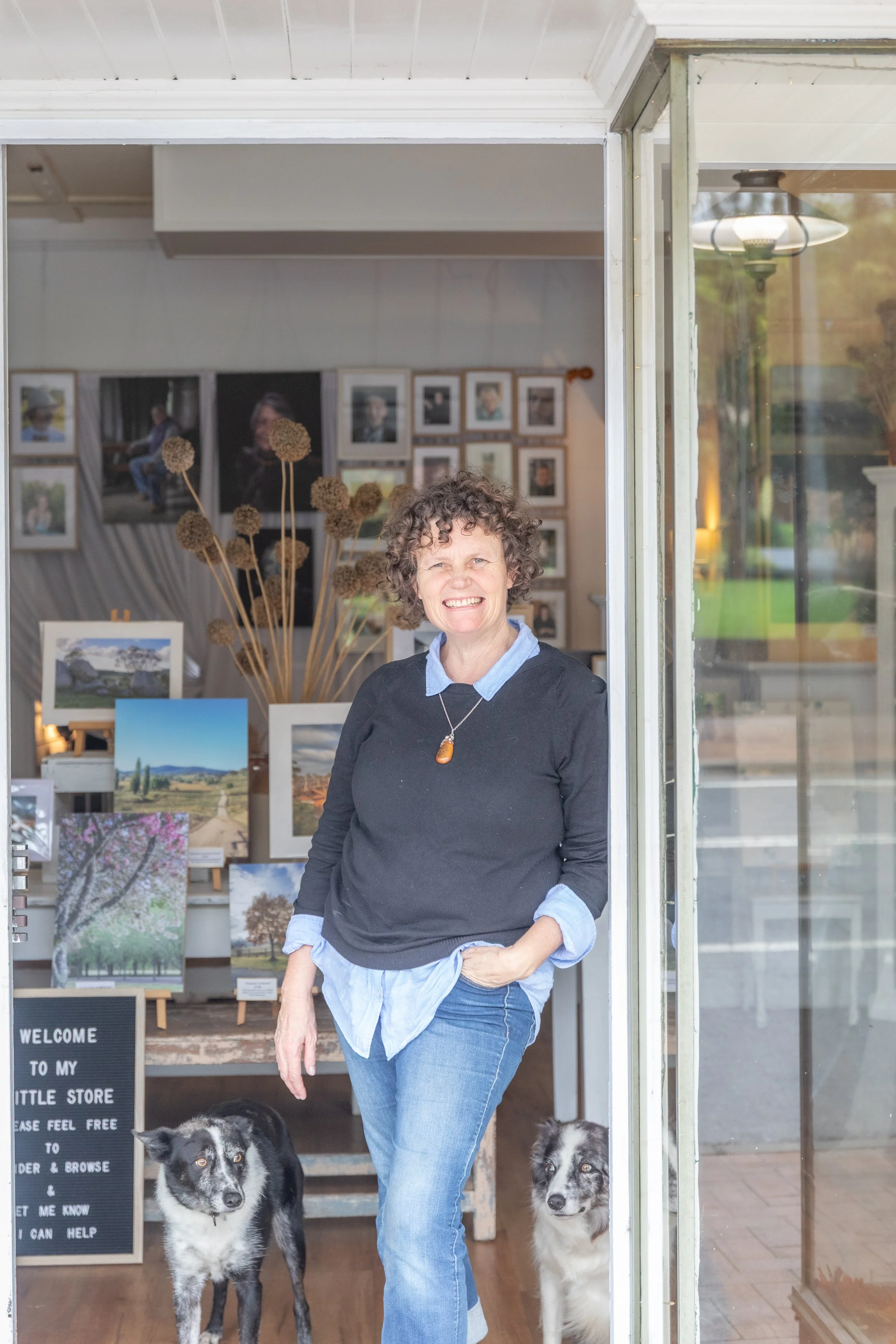 A woman with curly hair and a black sweater smiling while standing in front of an art gallery with two dogs, one on each side of her, inside the gallery with framed pictures and landscape photos visible in the background.