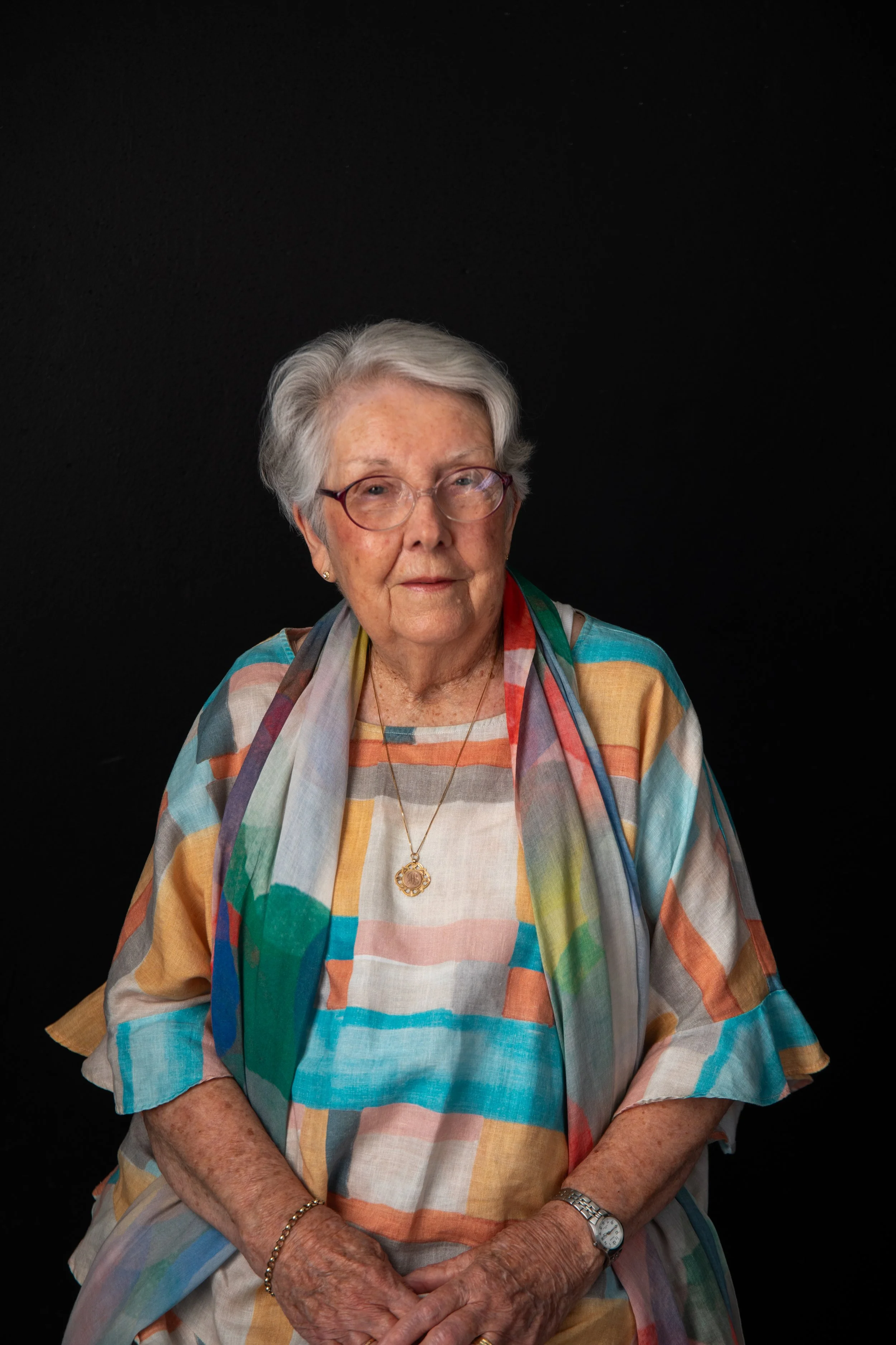 An elderly woman with short gray hair, wearing glasses, a colorful checkered dress, a multi-colored scarf, a wristwatch, and jewelry, sitting against a black background.