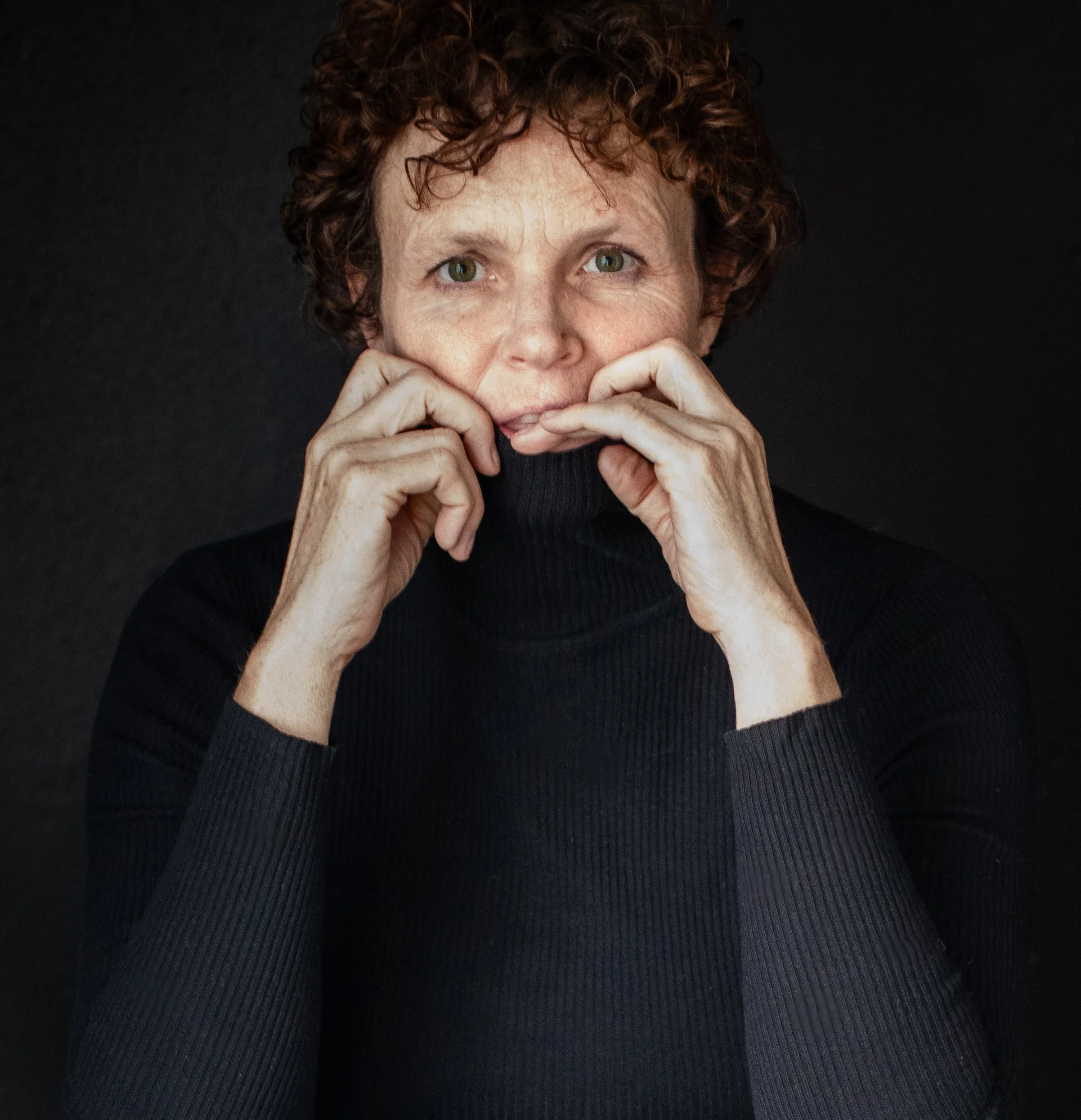 A woman with short, curly red hair wearing a black turtleneck, holding her hands close to her face against a black background.