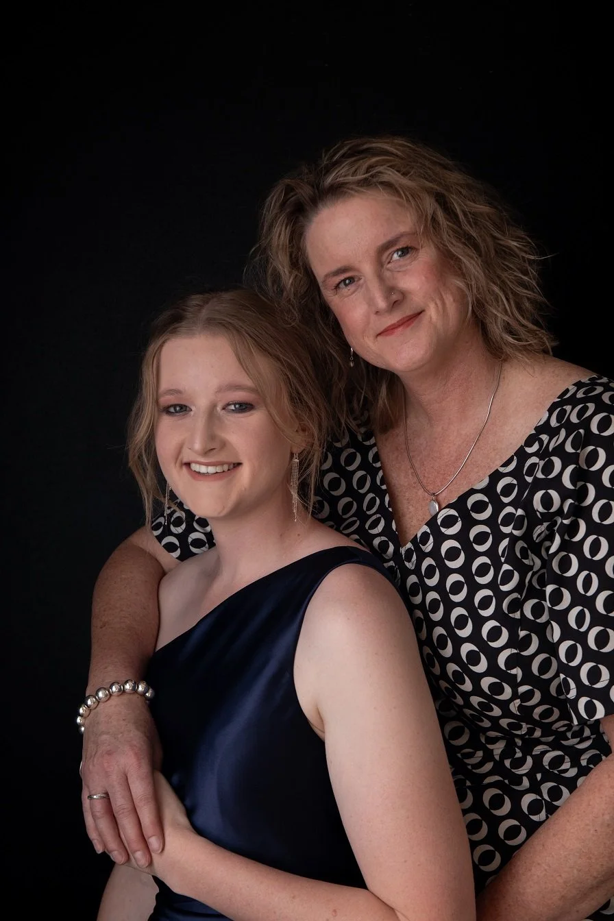 A woman and a young girl smiling and posing together against a black background. The woman is wearing a black and white patterned blouse, and the girl is in a navy blue dress with pearl jewelry.
