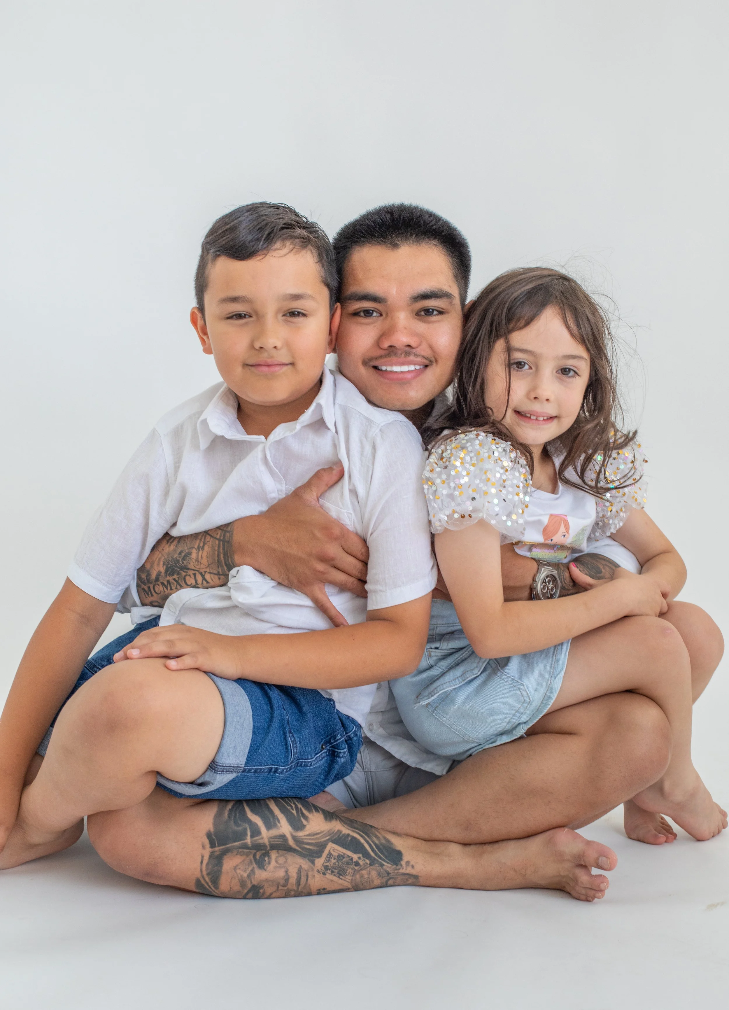 A man with tattoos and two children sitting on a plain white background, smiling and hugging each other.