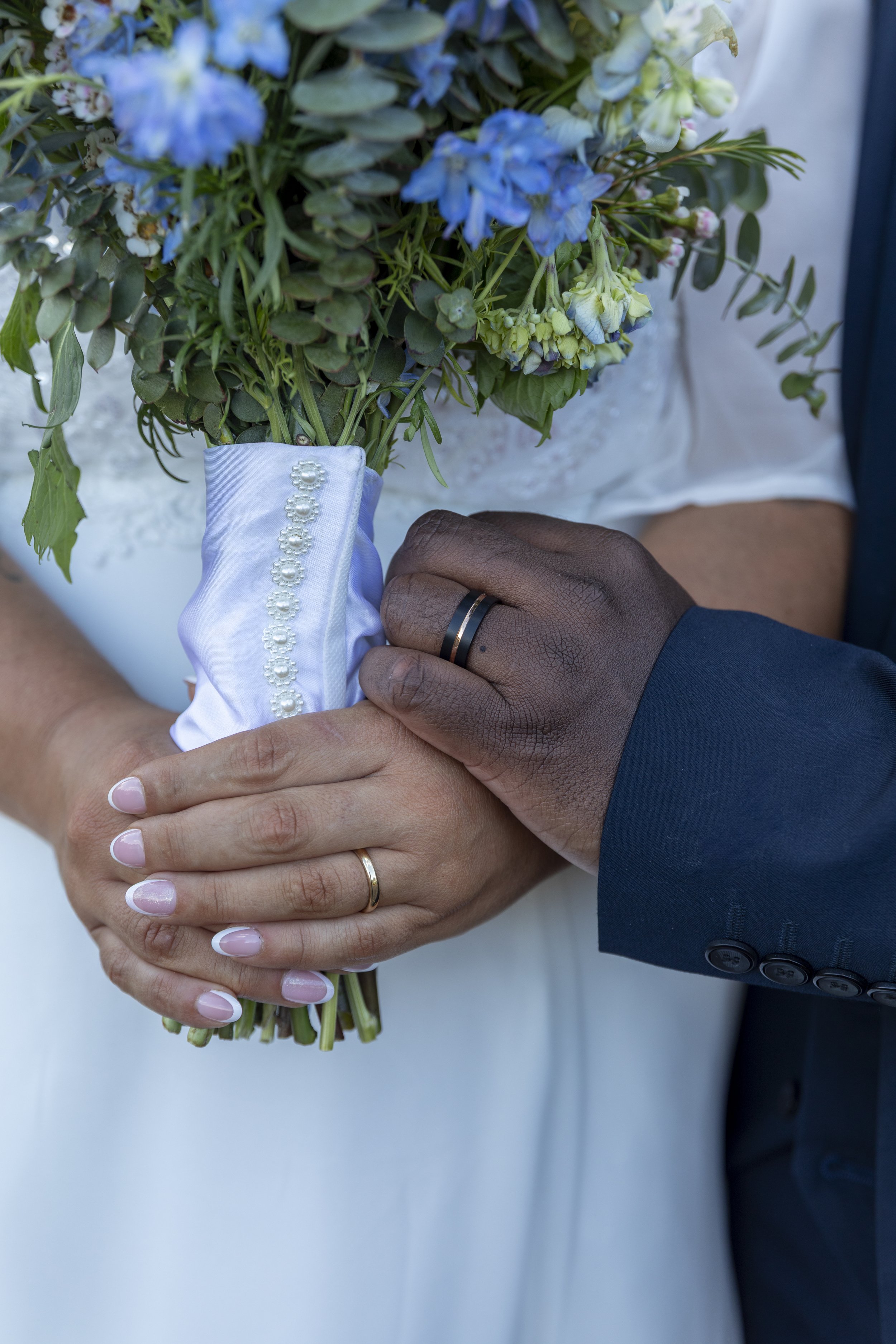 Close-up of a bride and groom holding a bouquet of flowers together, both wearing wedding rings. The bride's hand is visible with a gold ring, and the groom's hand with a black ring. The bride's bouquet contains blue and white flowers, and she is wea
