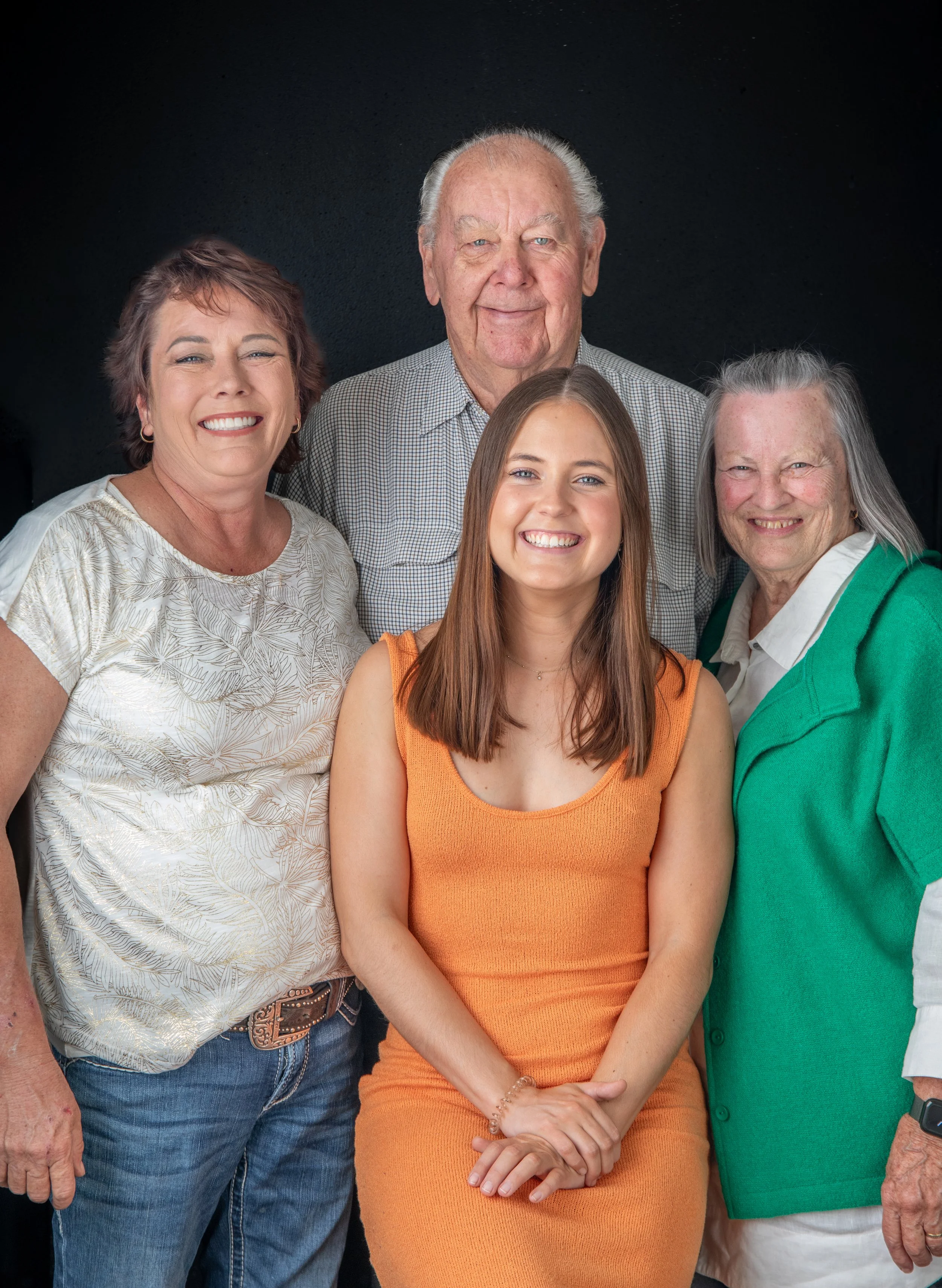 A group of five people, three women and two men, smiling against a dark background, with a young woman in an orange dress in front.