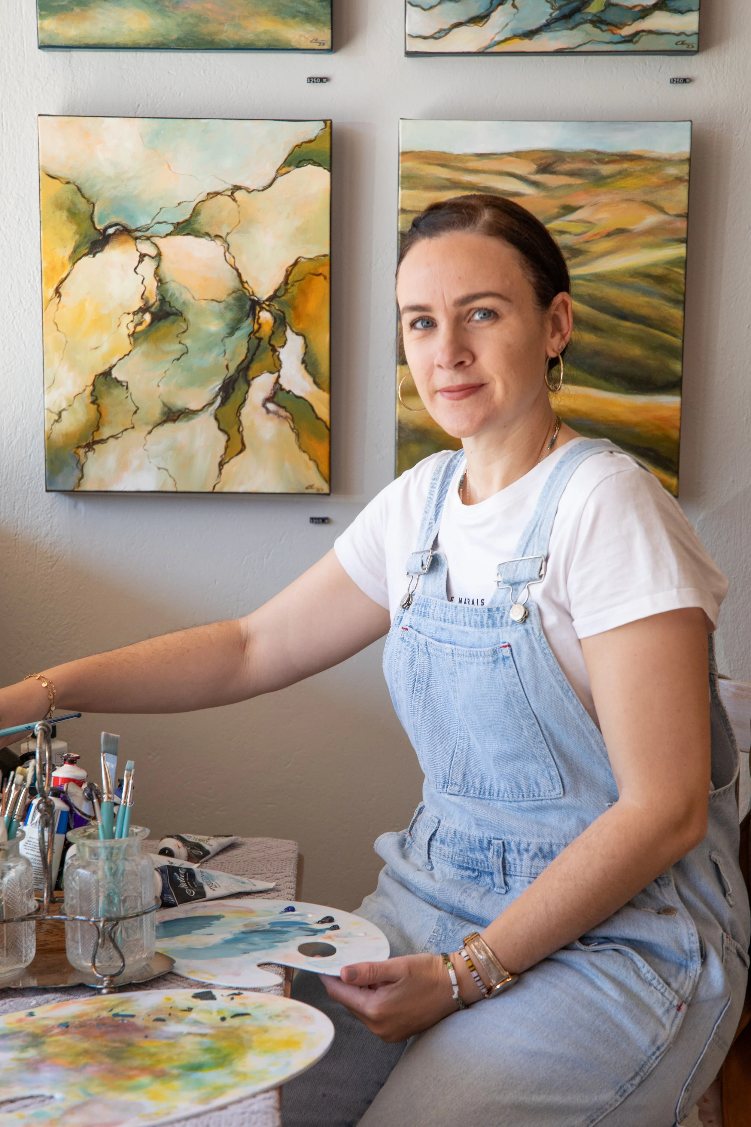 A woman sitting at an art table with painting supplies, holding a palette, in front of abstract landscape paintings on the wall.