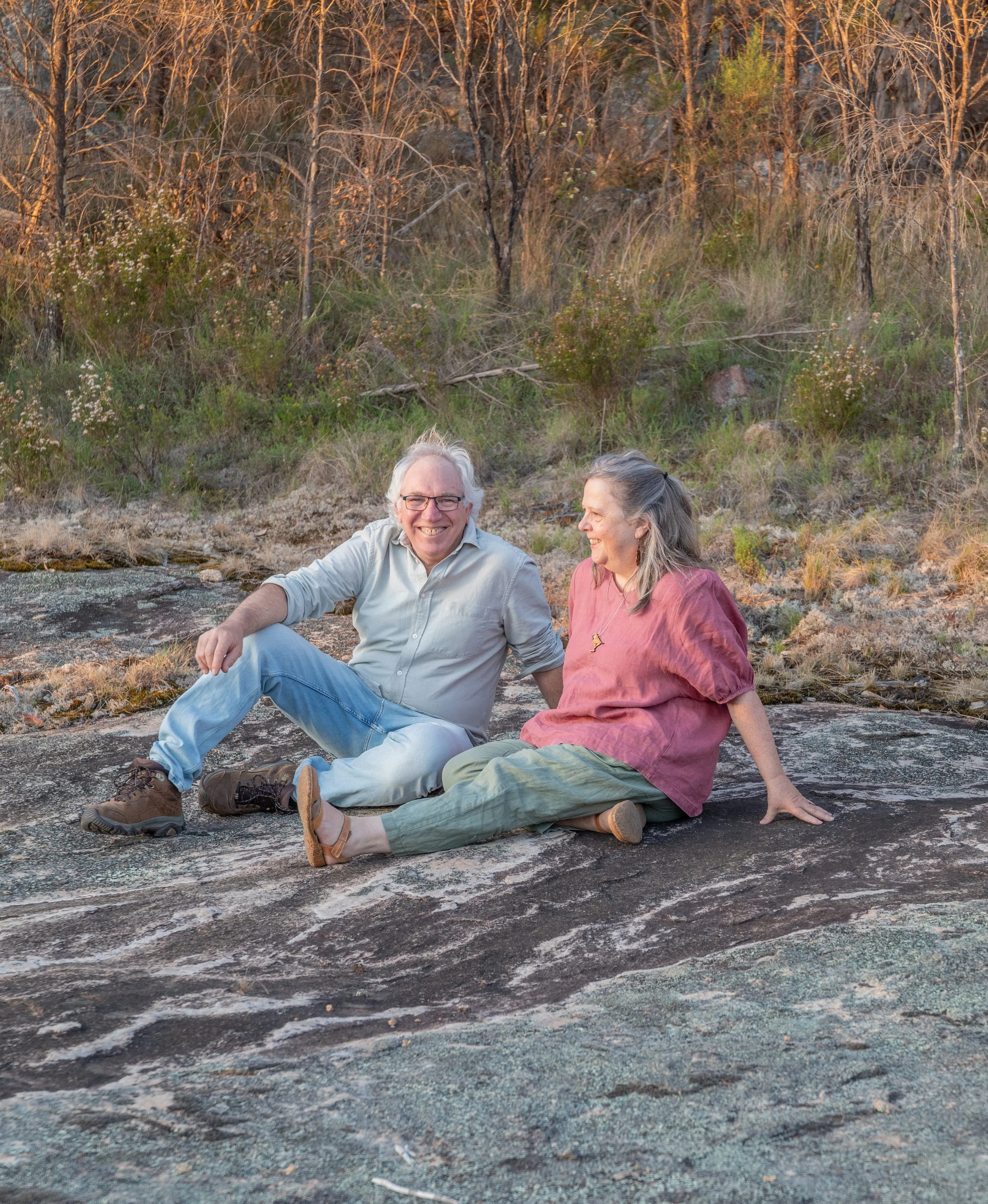 An elderly man and woman sitting on a large rock outdoors, smiling and enjoying each other's company during sunset or sunrise in a natural setting with trees and shrubs in the background.