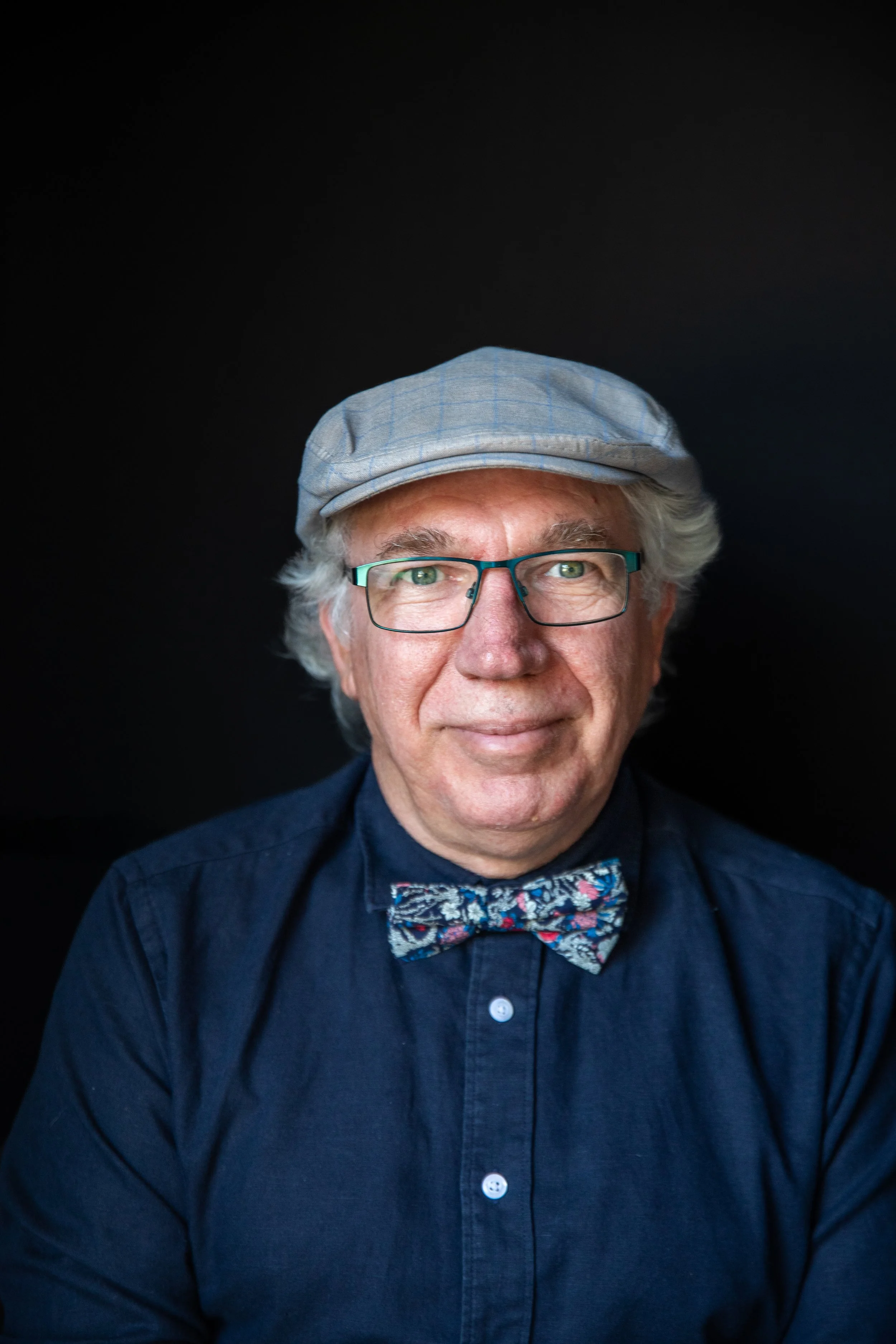 Portrait of an elderly man with glasses, a gray flat cap, a dark shirt, and a colorful bow tie against a black background.