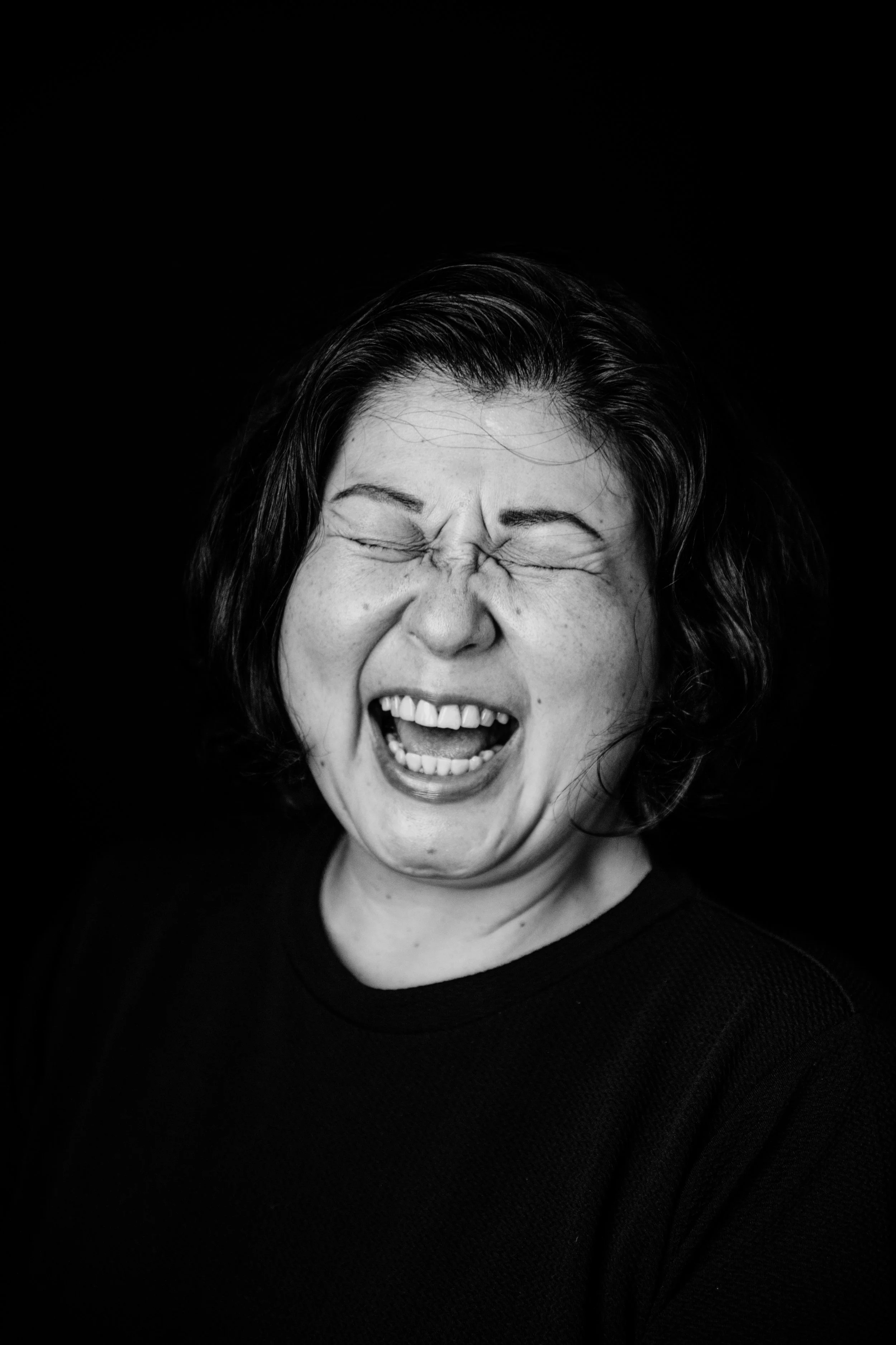 Black and white photo of a woman smiling broadly with eyes closed, showing joy, wearing a dark top, against a black background.