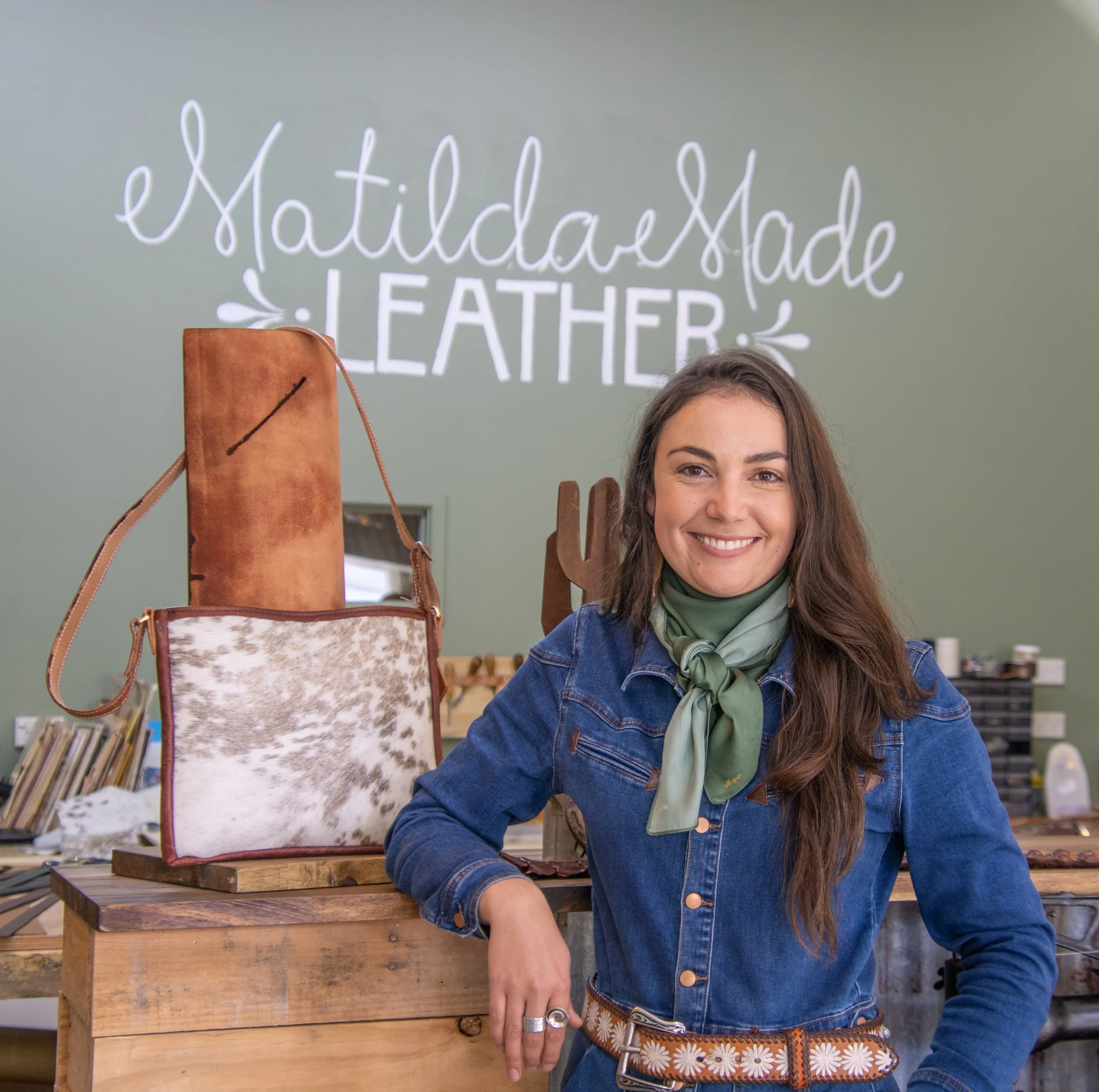 Woman smiling at the camera in a leather workshop with a green wall background, handwritten sign saying 'MatildaMade Leather,' and leather bags on display.