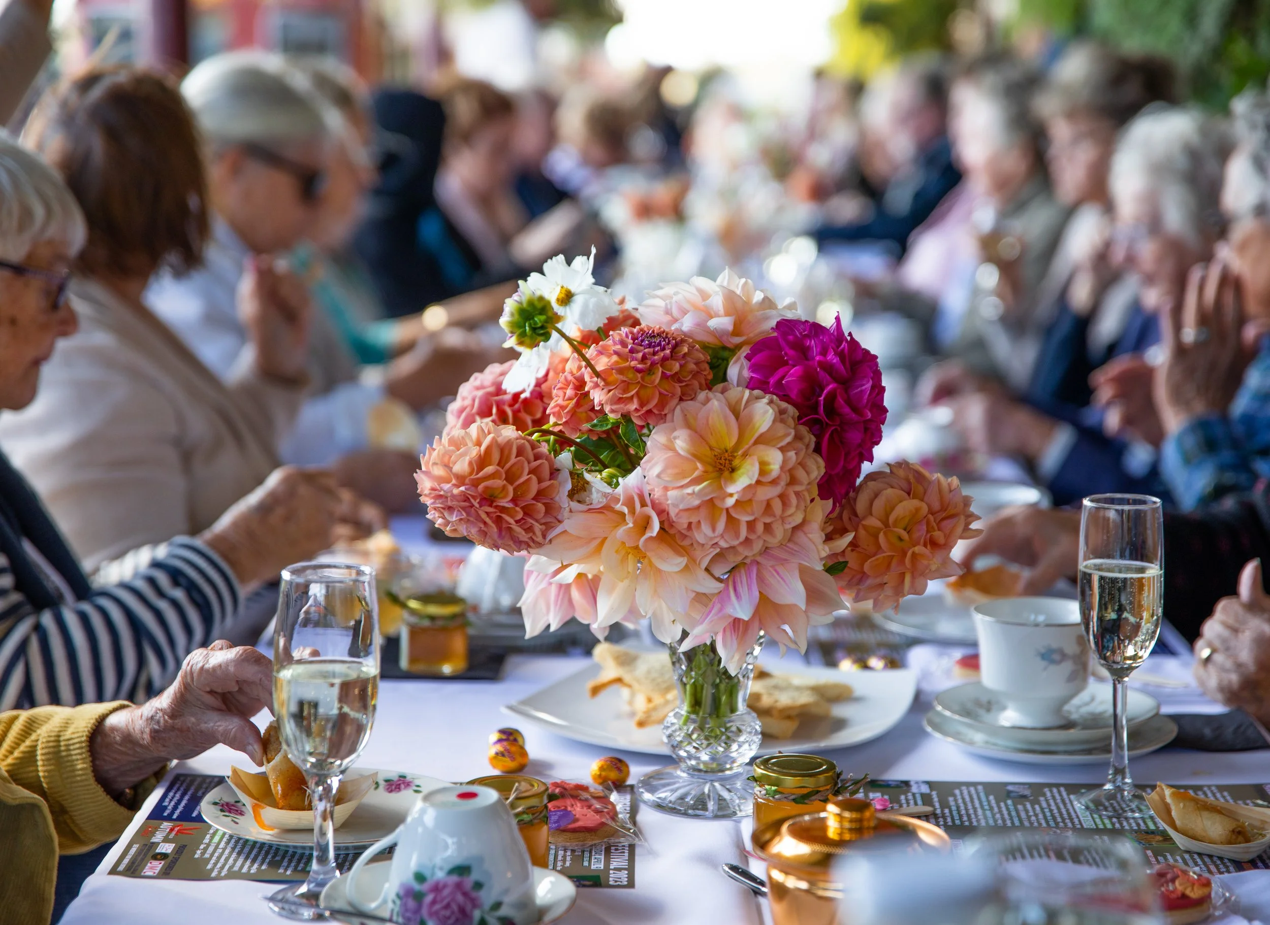 A group of elderly people gathered around a long table for a meal, with a large bouquet of pink, peach, and white flowers as a centerpiece.
