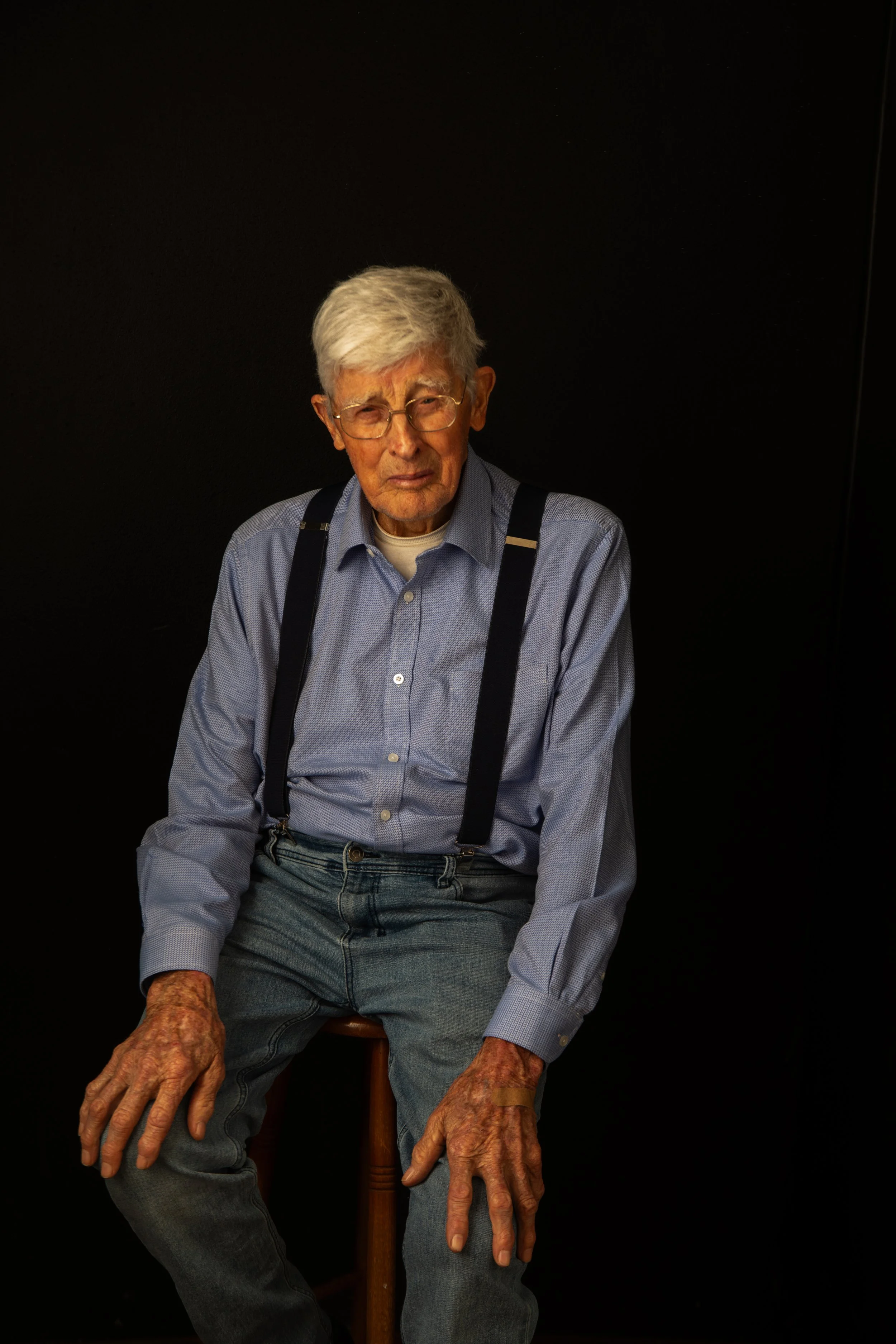 An elderly man with gray hair and glasses, wearing a blue button-up shirt and suspenders, sitting on a wooden stool against a black background.