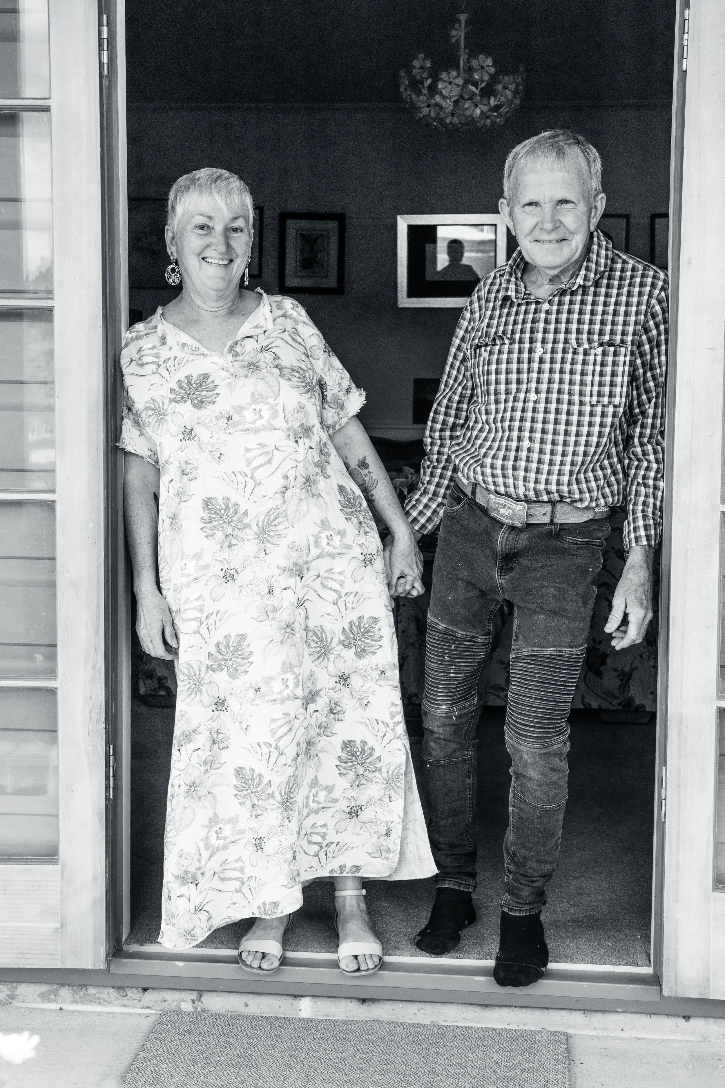 A smiling woman and man holding hands, standing in a doorway, inside a home with framed pictures on the wall.
