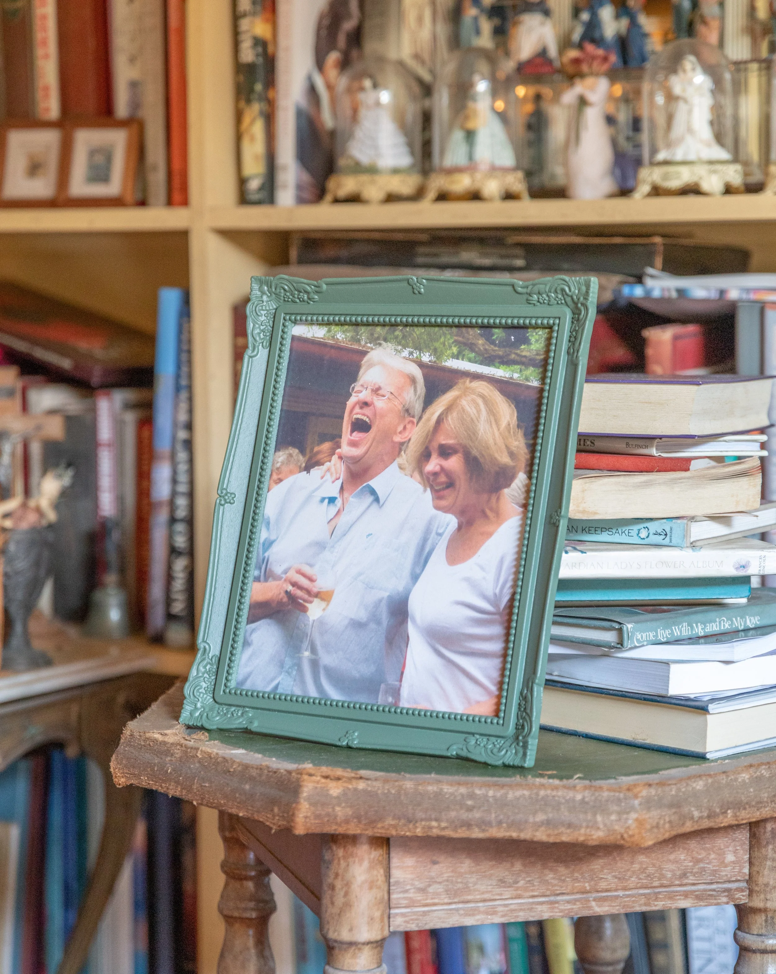 A framed photograph of an elderly man and woman laughing and enjoying drinks at an outdoor gathering, placed on a small wooden table surrounded by books and decorative figurines.