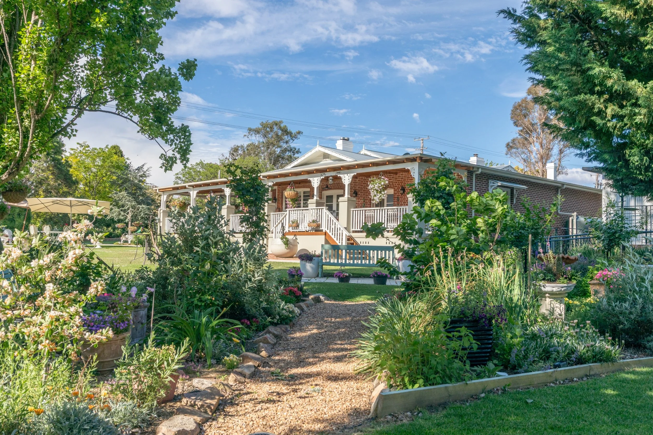 A brick house with a white porch, surrounded by a garden with various plants, flowers, and trees, under a partly cloudy sky.