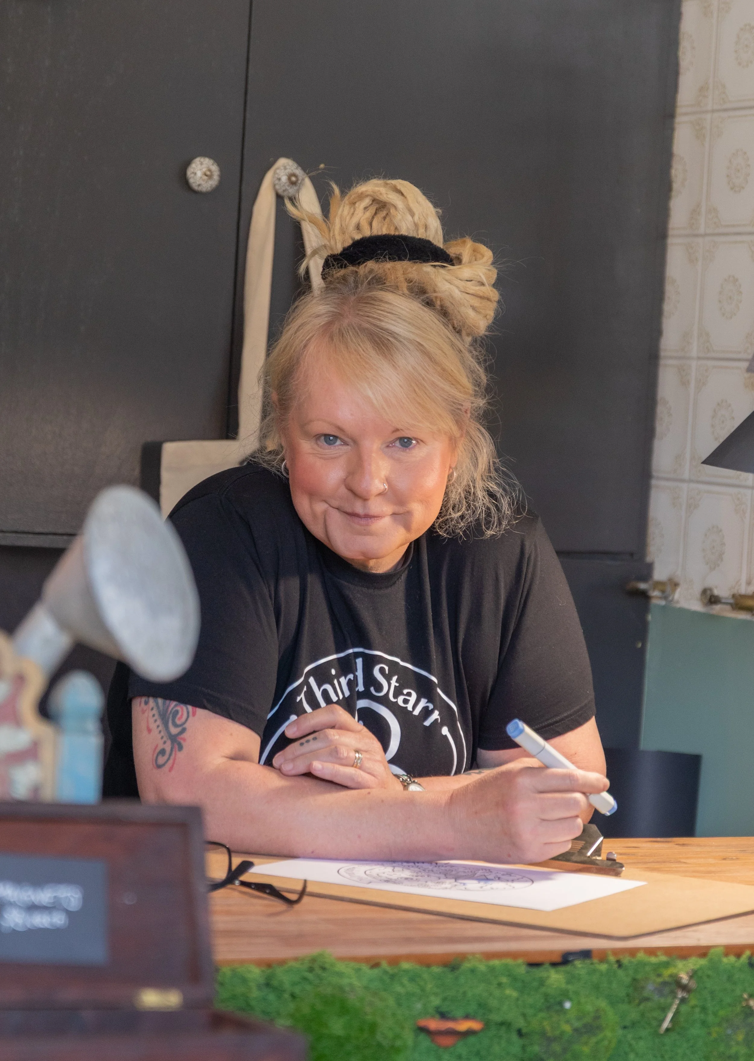 A woman with blonde hair, tied up in a large bun with a black scrunchie, sitting at a table with a pen in hand, looking at the camera and smiling. She is wearing a black T-shirt with a circular logo that says 'Third Star'. There are some drawing tool