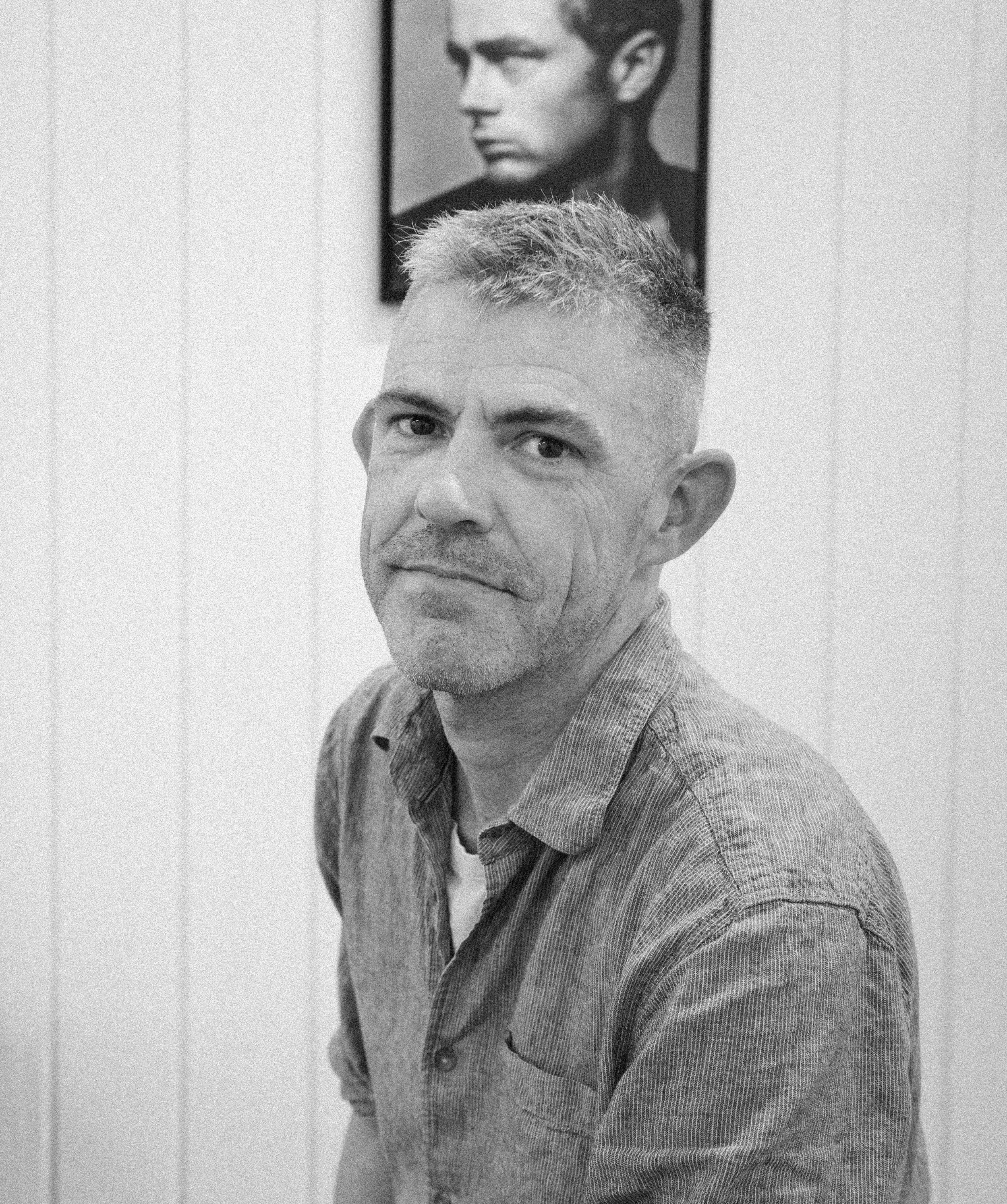 Black and white photo of a middle-aged man with short hair wearing a collared shirt, sitting indoors with a framed portrait of a young man in the background.