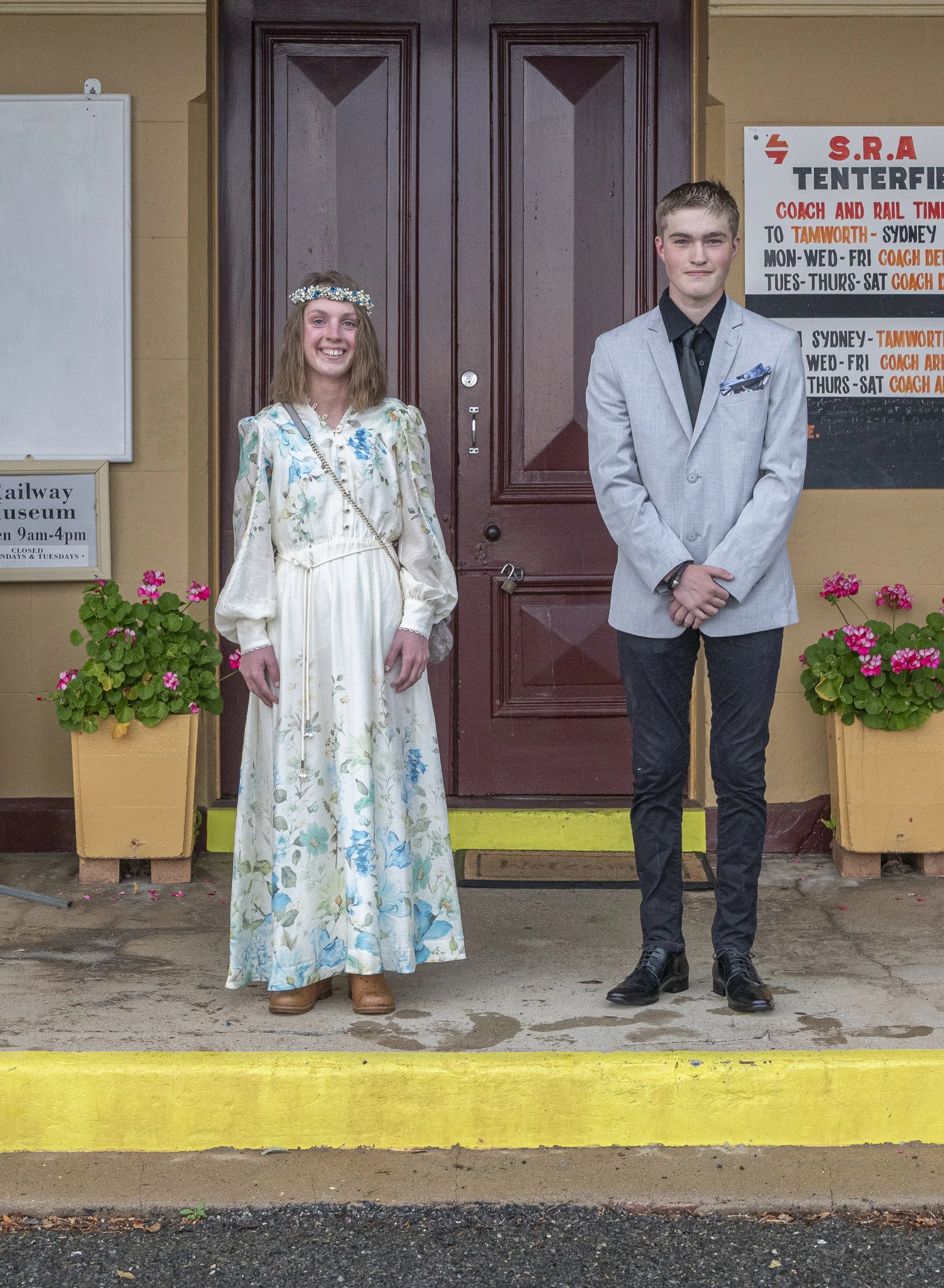A young girl and a young boy standing outside in front of a wooden door. The girl is wearing a long, floral dress and a flower crown, smiling. The boy is dressed in a gray suit with a black shirt and tie, with his hands clasped in front of him. There
