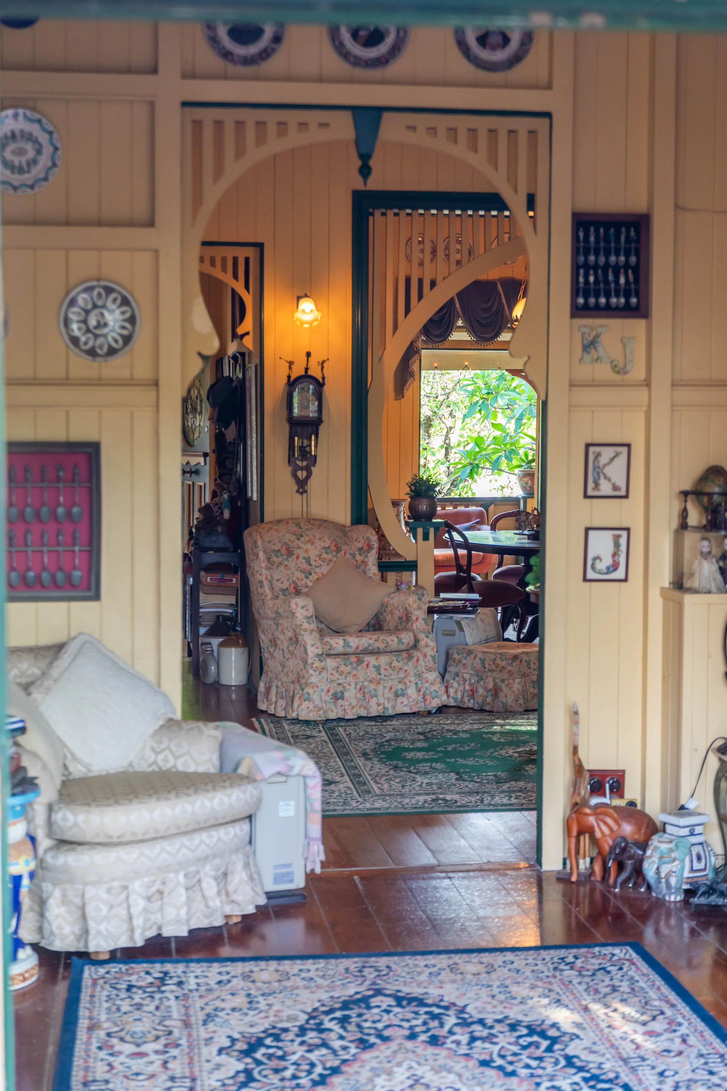 View of a cozy living room with vintage furniture, floral patterns, a green rug, and a bright window with lush greenery outside.