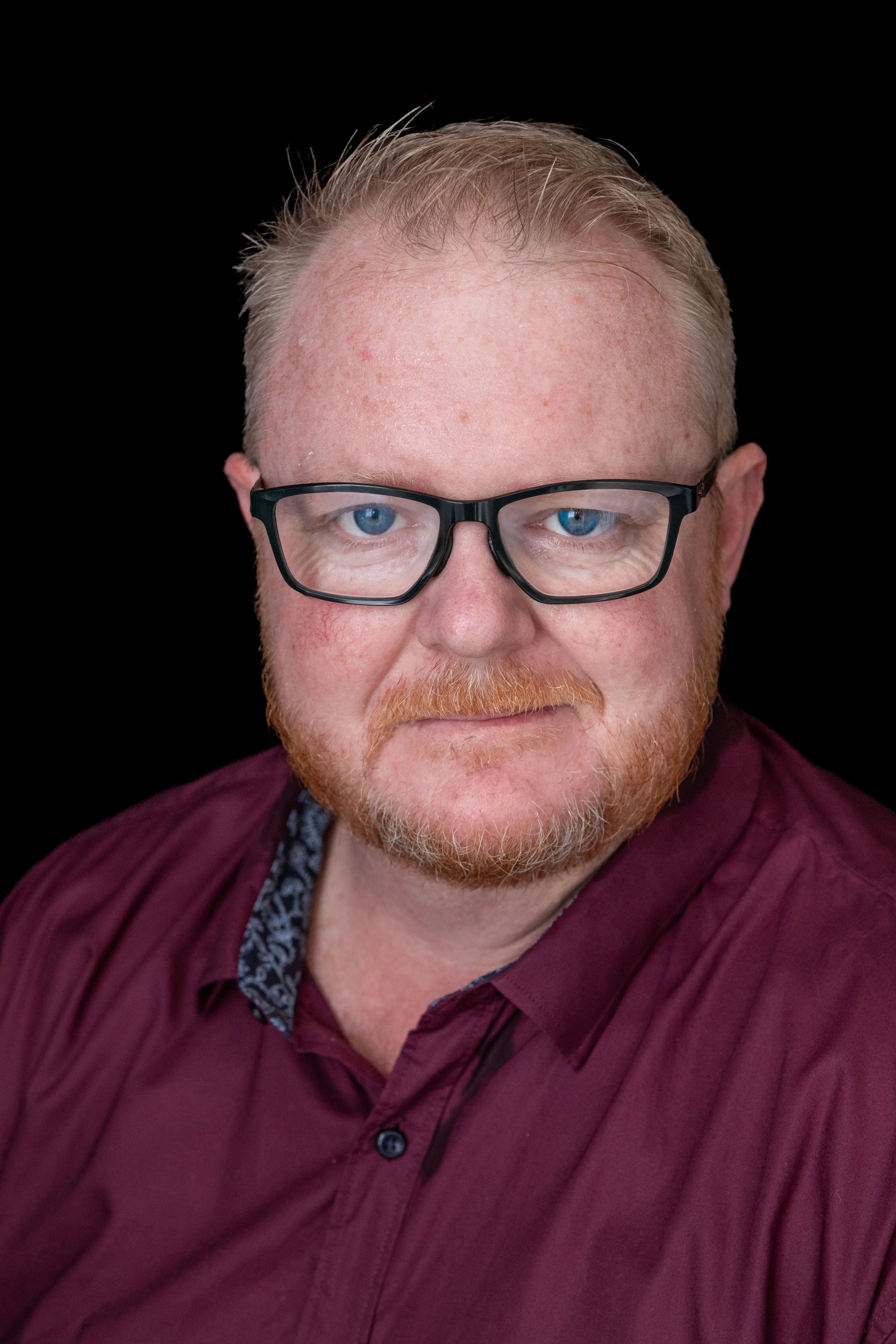 A man with red hair, beard, and glasses wearing a maroon shirt against a black background.