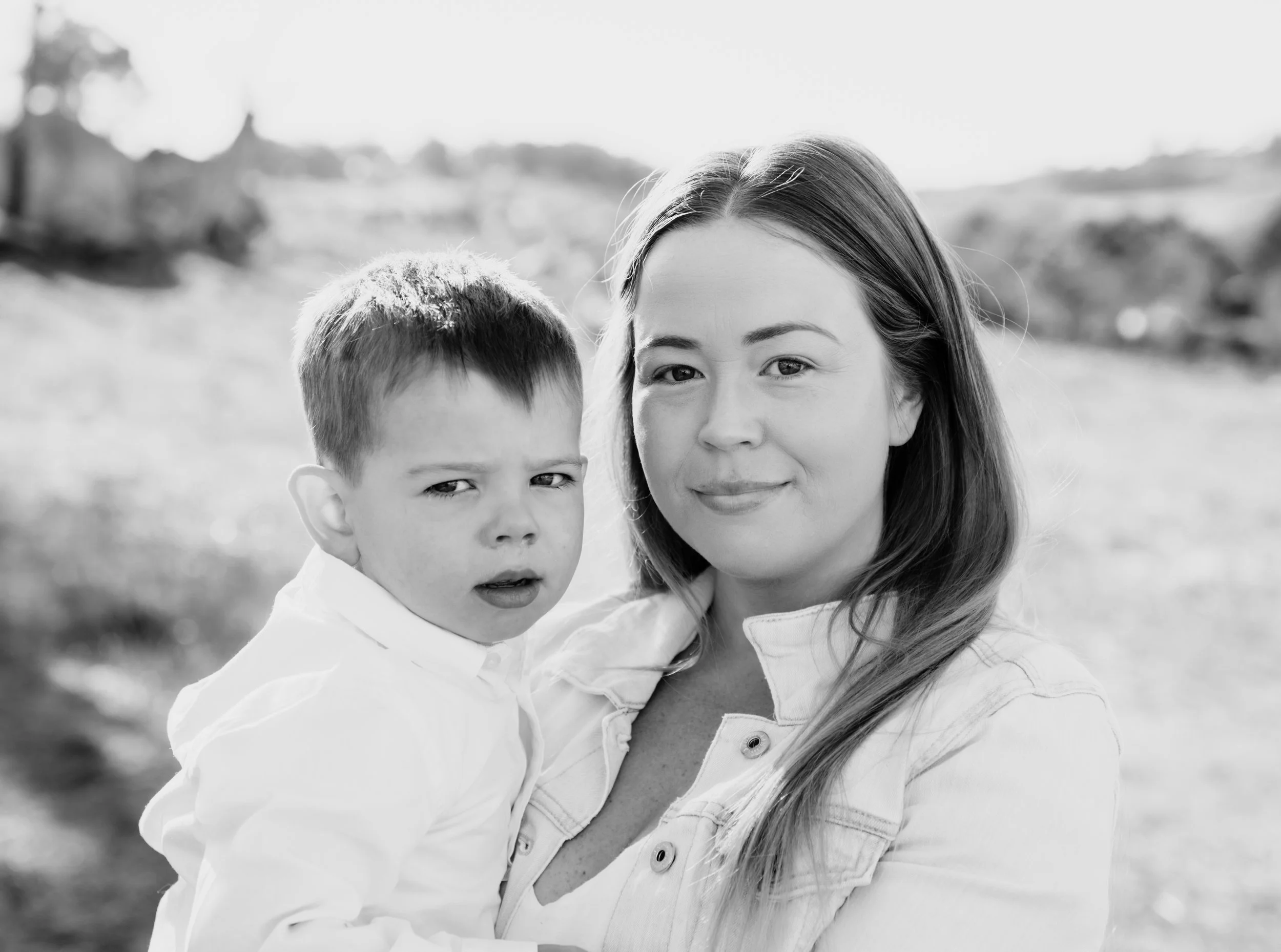 A woman holding a young boy outdoors with blurred trees and landscape in the background, black and white photo.