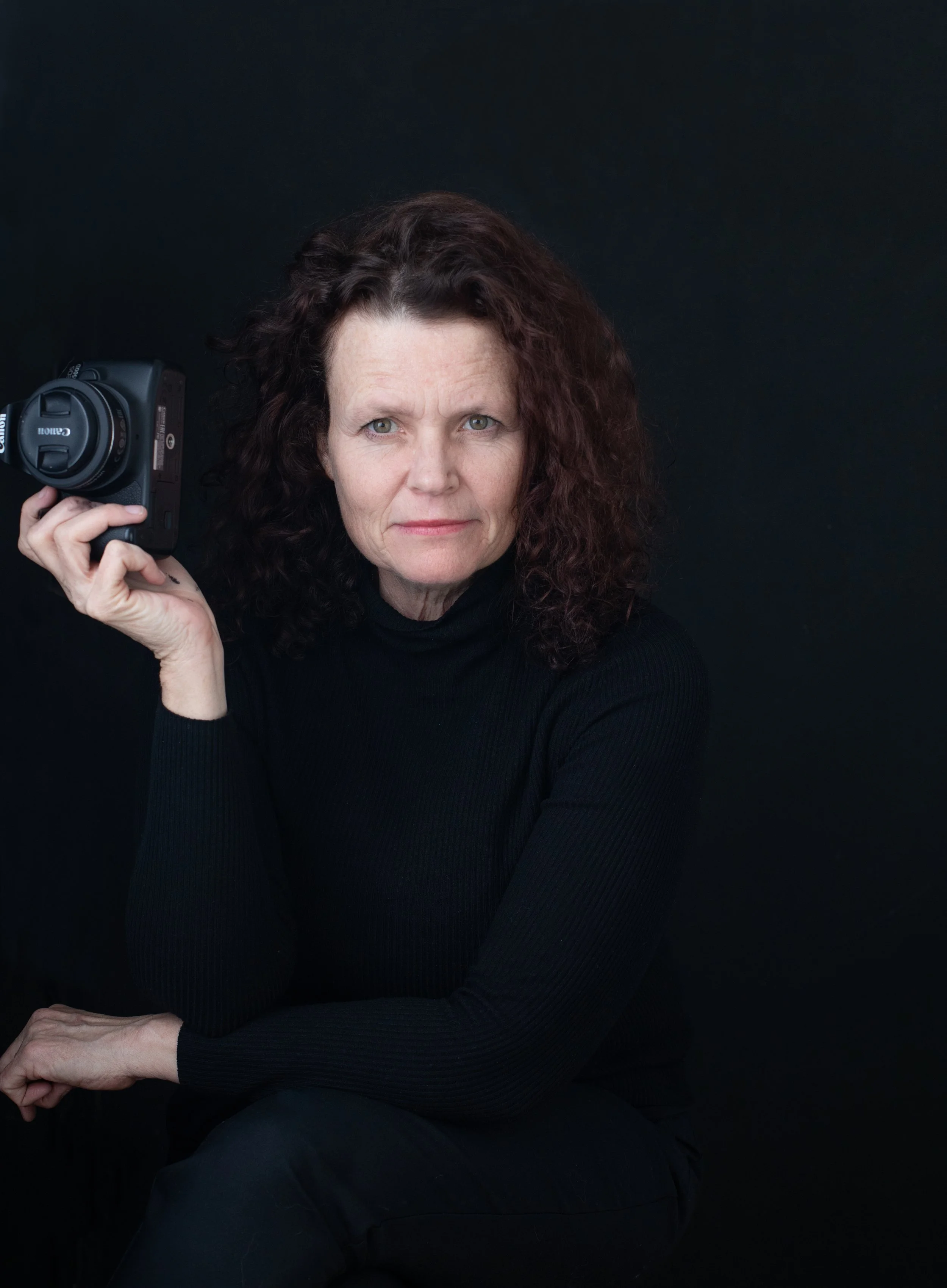 A woman with curly dark hair wearing a black turtleneck, holding a Canon DSLR camera against a black background.