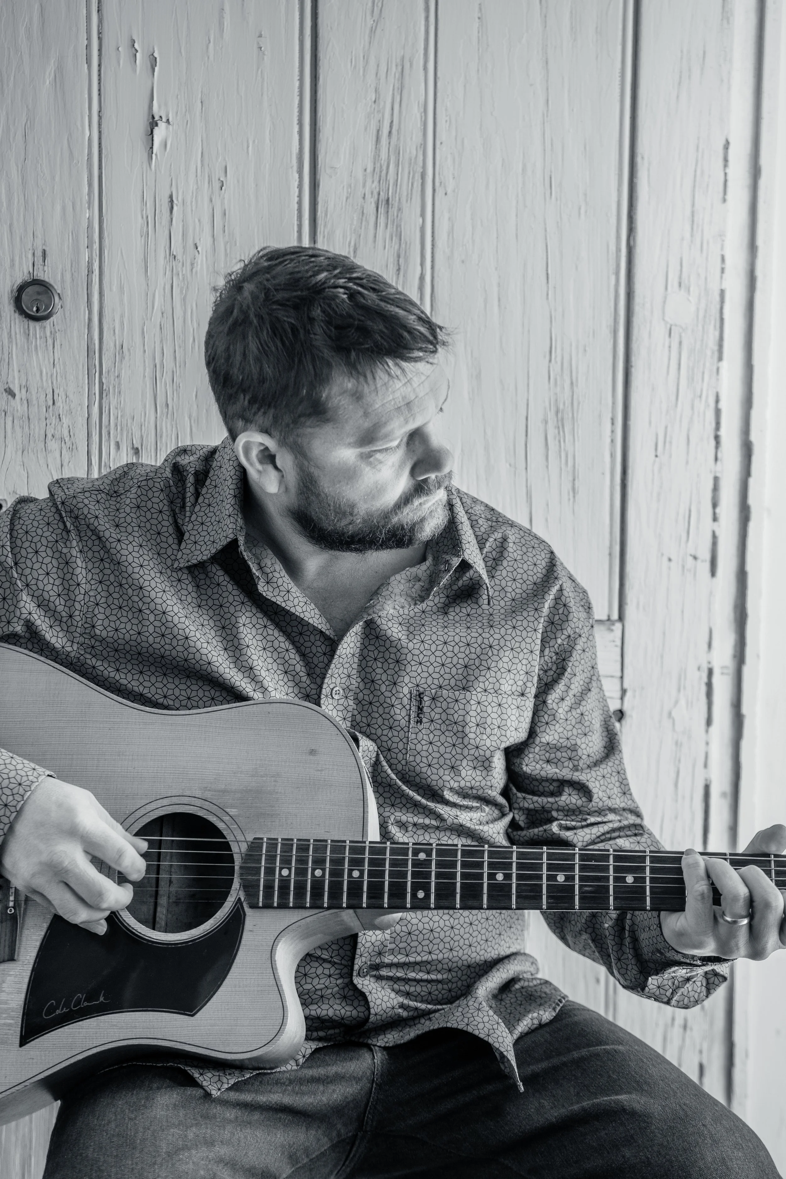 A man with short hair and a beard sitting against a wooden wall, playing an acoustic guitar, looking to his right.