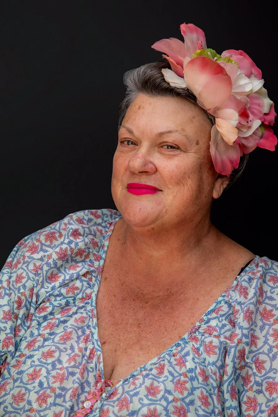 Portrait of an older woman with short gray hair, wearing bright pink lipstick, a large pink flower headpiece, and a floral patterned blouse against a black background.