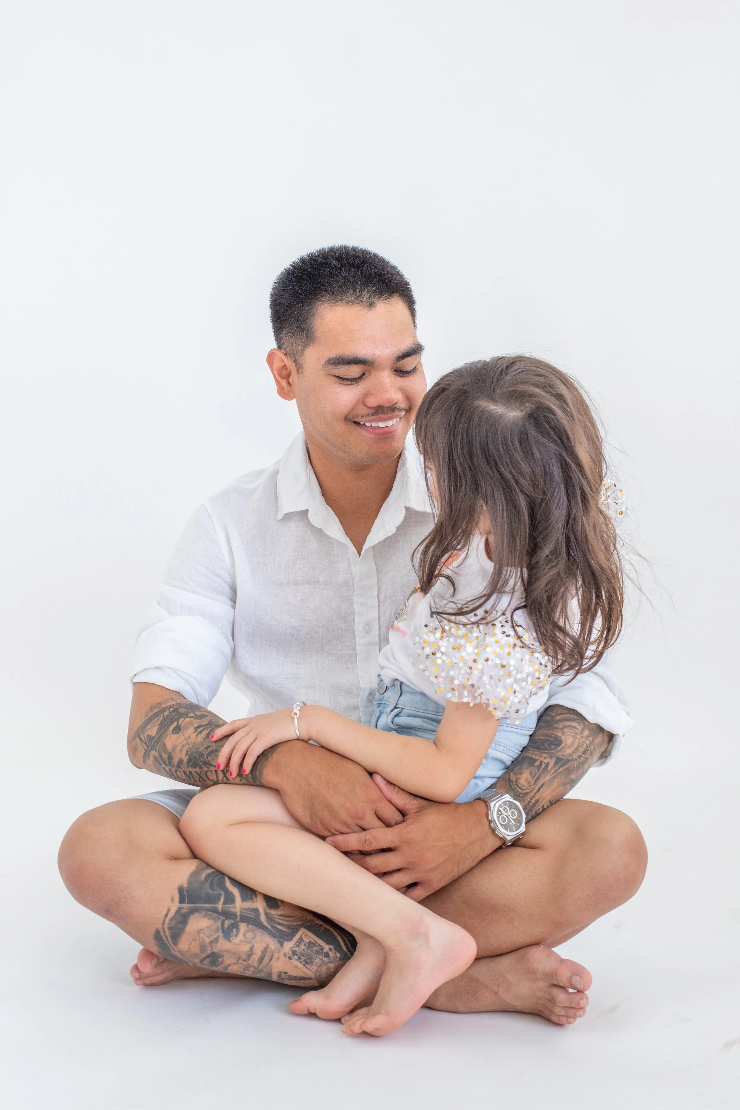 A man and young girl sitting on the floor, hugging and looking at each other with smiles. The man has tattoos on his arms and legs, and wears a white shirt and a watch. The girl has long brown hair, is wearing a white top with colorful beads and blue