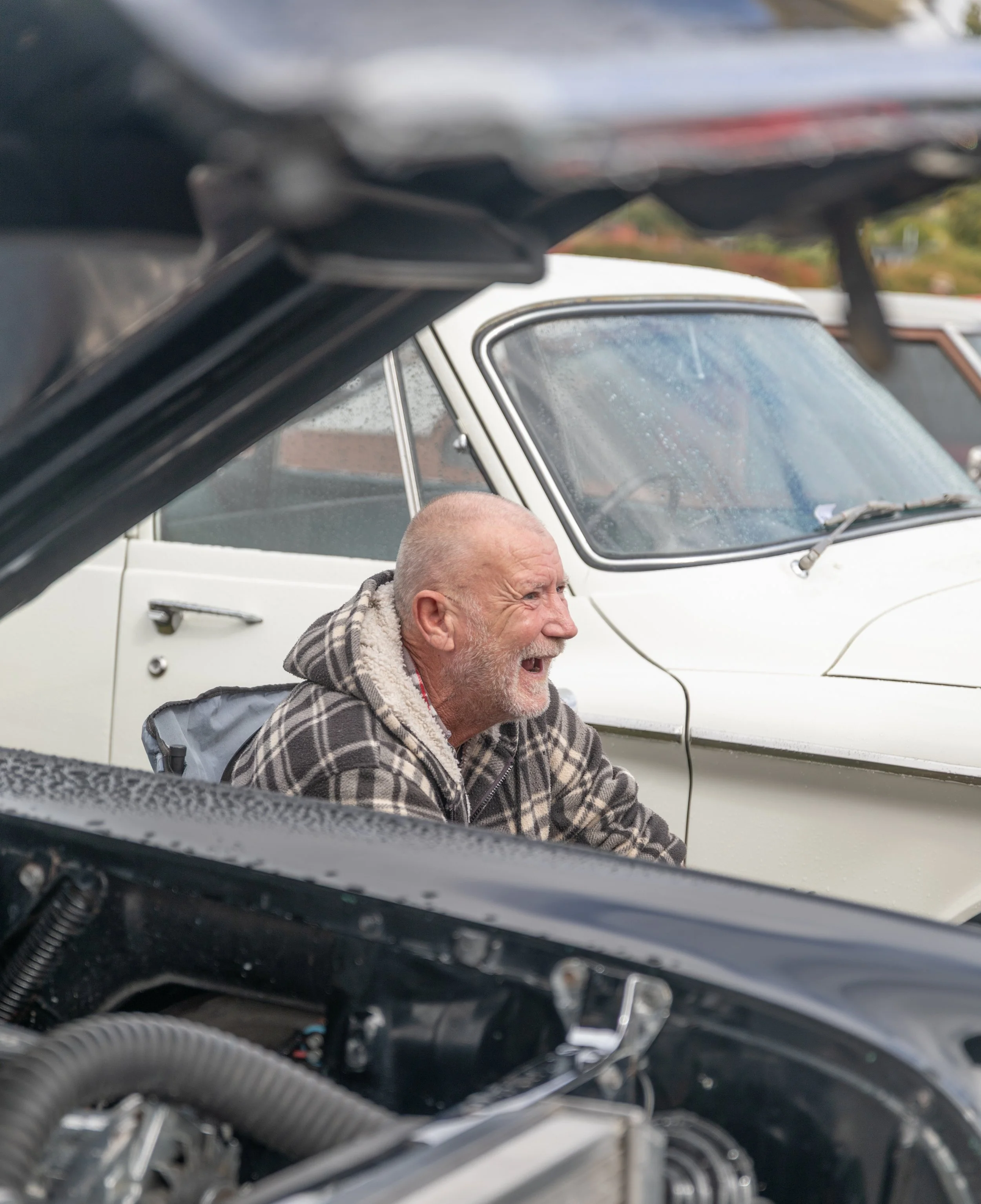 An elderly man with a gray beard and shaved head, wearing a checkered hoodie, is smiling and crouching next to a classic white car and a black vehicle, possibly engaging in car repair or maintenance outdoors.