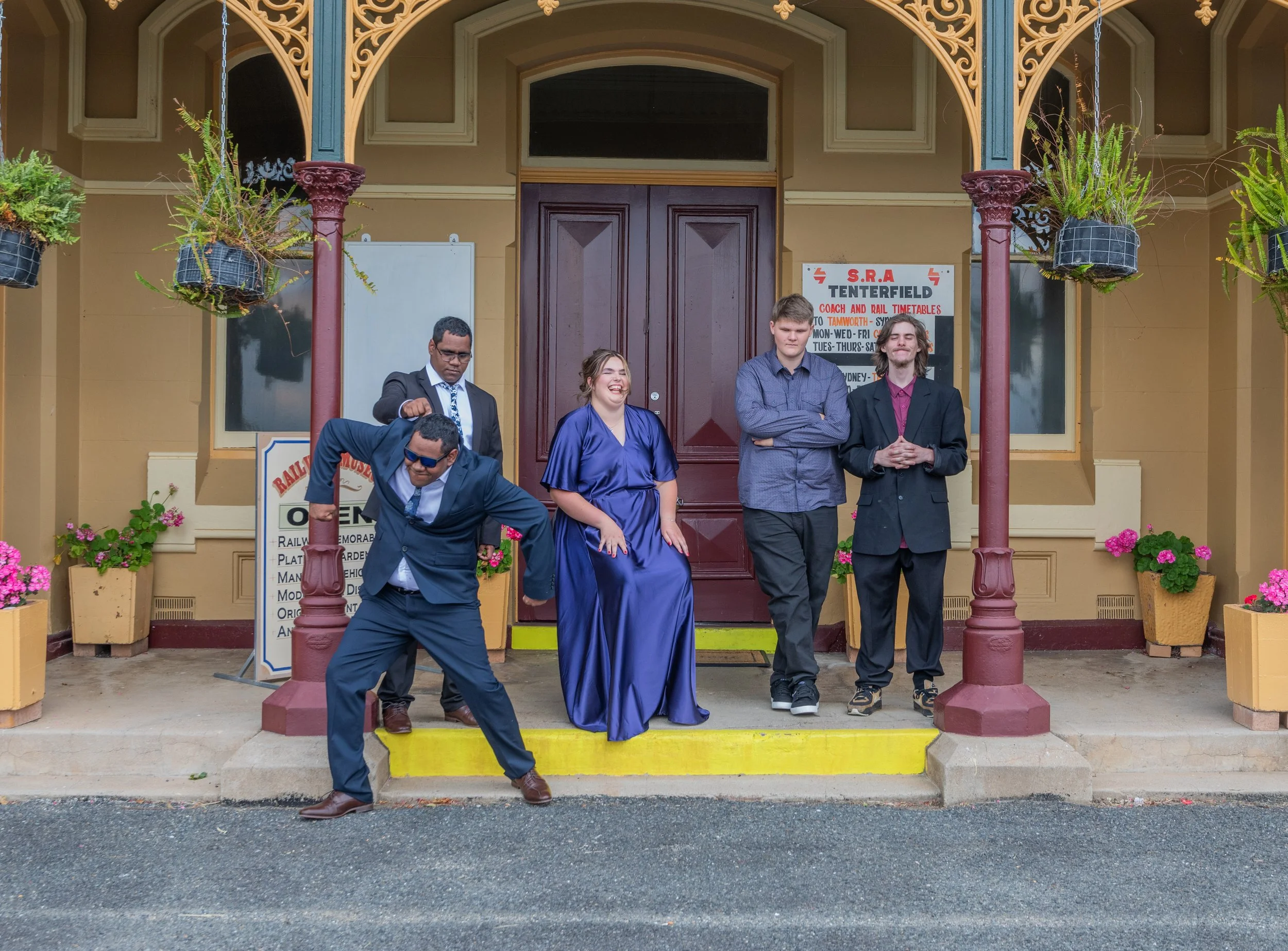 Five people standing and sitting on the steps of a vintage building with a sign that says 'S.R.A TENTERFIELD Coach and Rail Timetables' and a whiteboard. The group includes four men dressed in suits and one woman in a blue satin dress, all smiling an