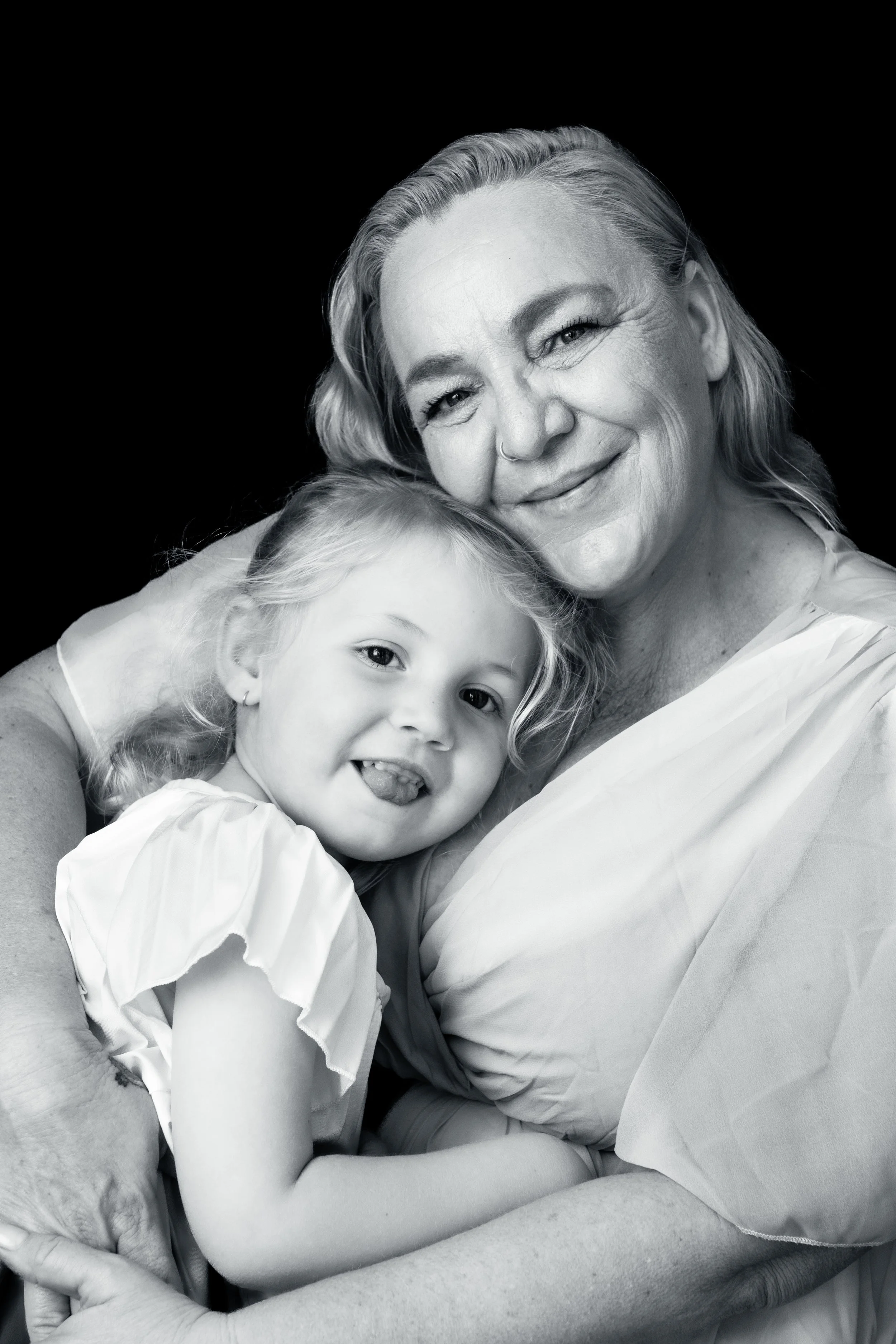 Black and white portrait of an older woman with a young girl, both smiling and hugging. The woman has wavy hair and a nose ring, the girl has curly hair and is showing her tongue.