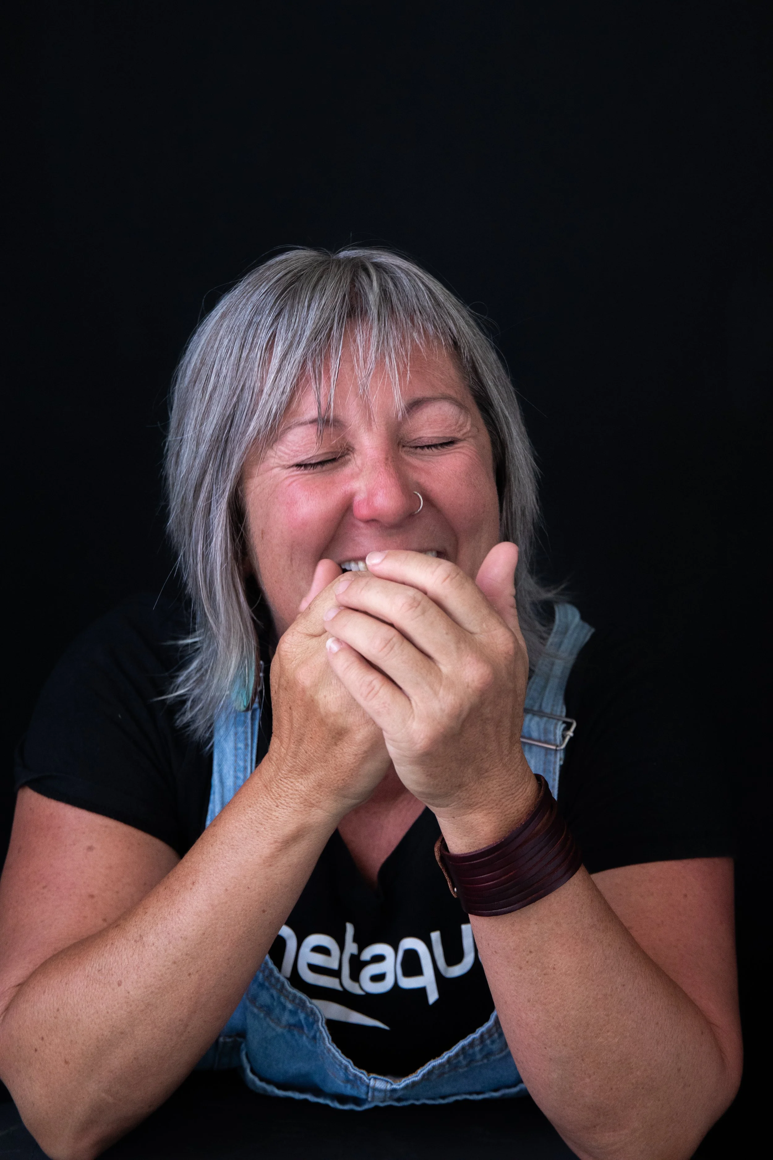 A woman with short gray hair smiling and covering her mouth with her hands against a black background.