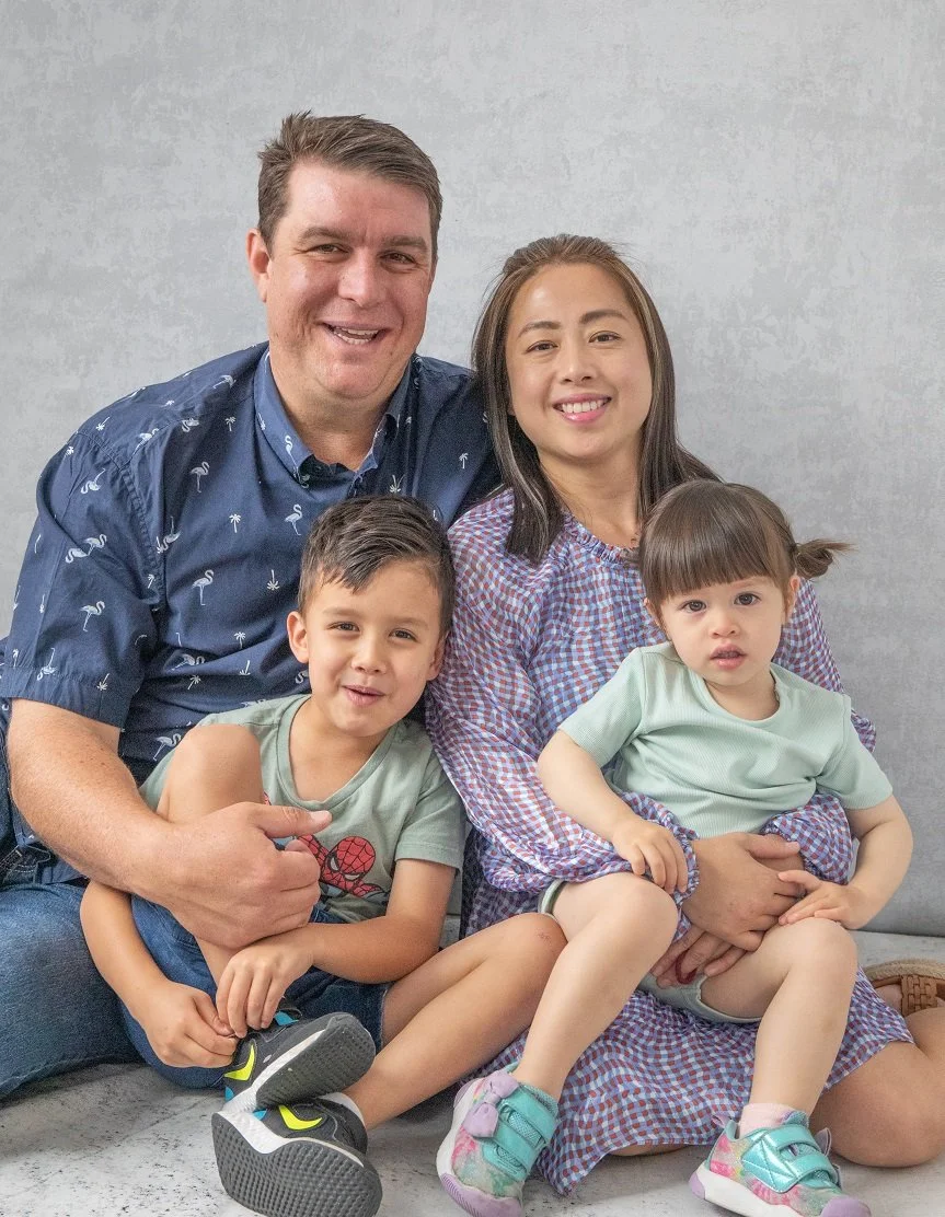 A family of four sitting on a gray couch, smiling at the camera. The father has short brown hair and is wearing a blue shirt with a palm tree pattern. The mother has long dark hair and is dressed in a patterned shirt. The young boy is wearing a gray 