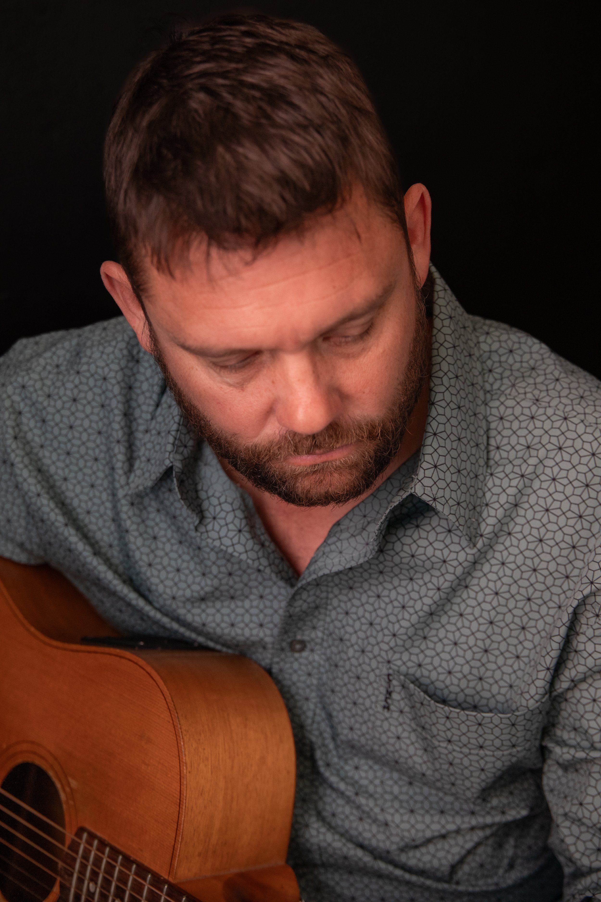 A man with short brown hair and beard wearing a patterned button-up shirt, looking down while playing an acoustic guitar.