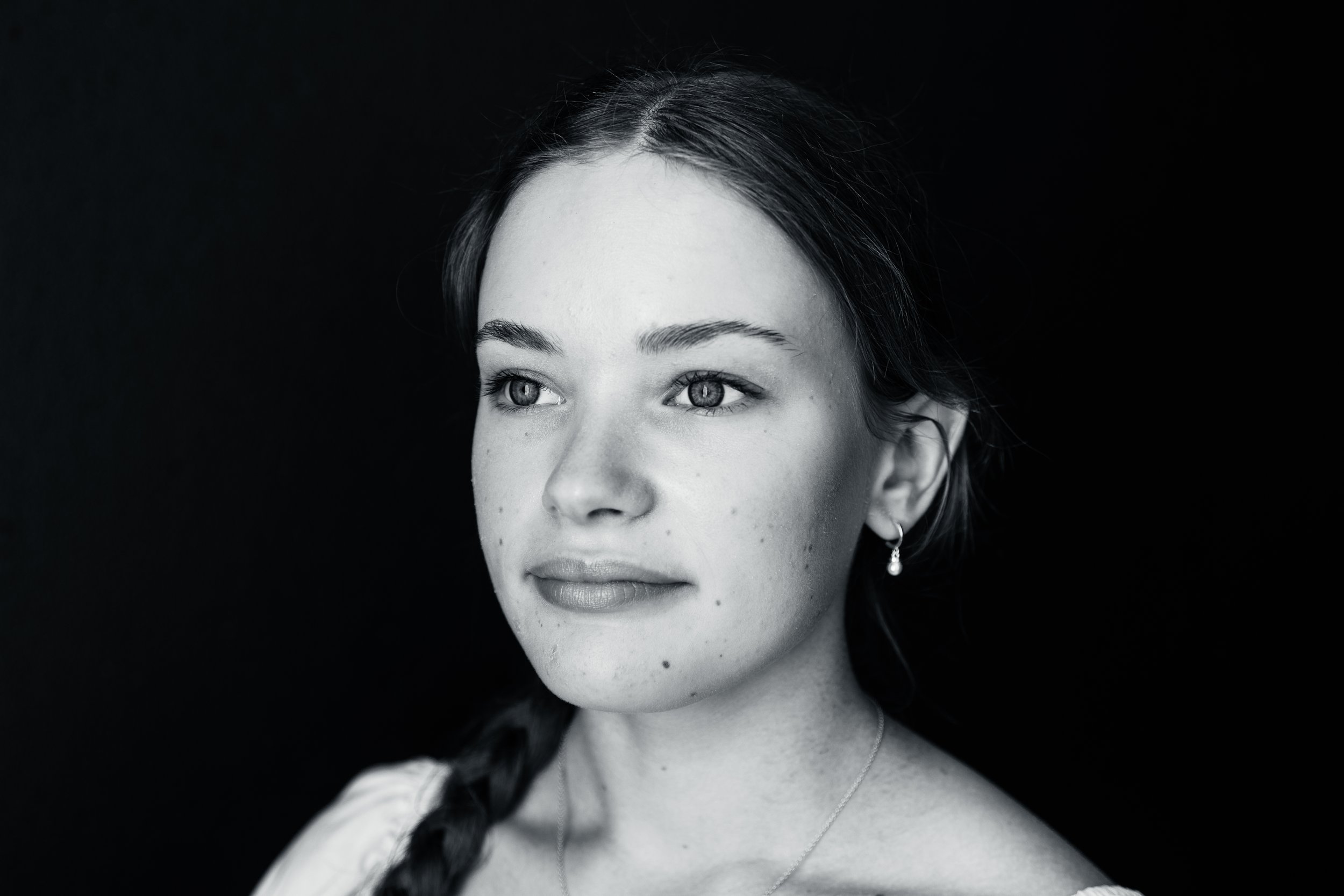 Black and white portrait of a young woman with light skin and dark hair styled in a braid, wearing earrings and a necklace, looking slightly to the side against a dark background.