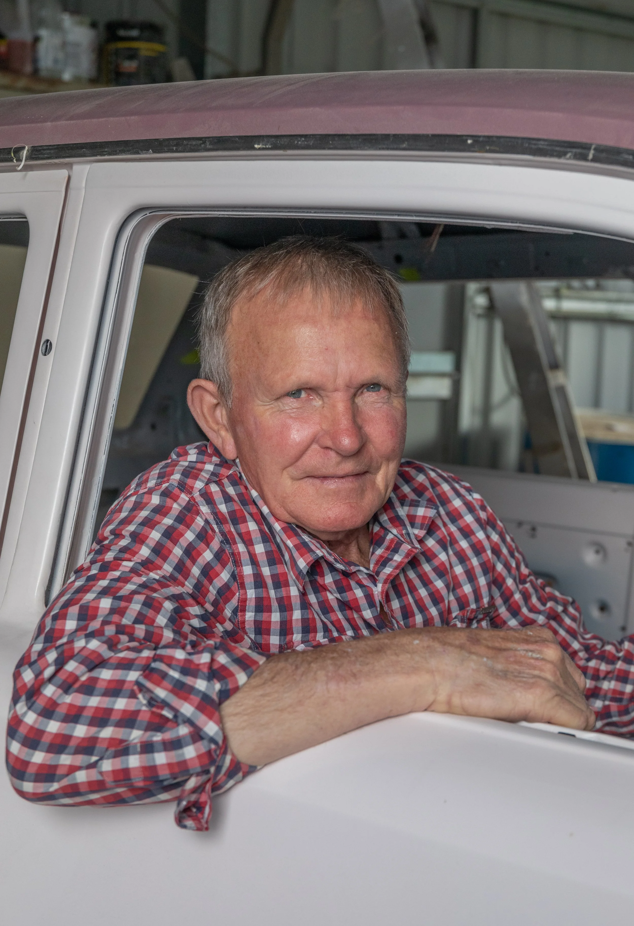 An older man with a checkered red and blue shirt sitting inside a vintage car, resting his arm on the window frame, looking at the camera.