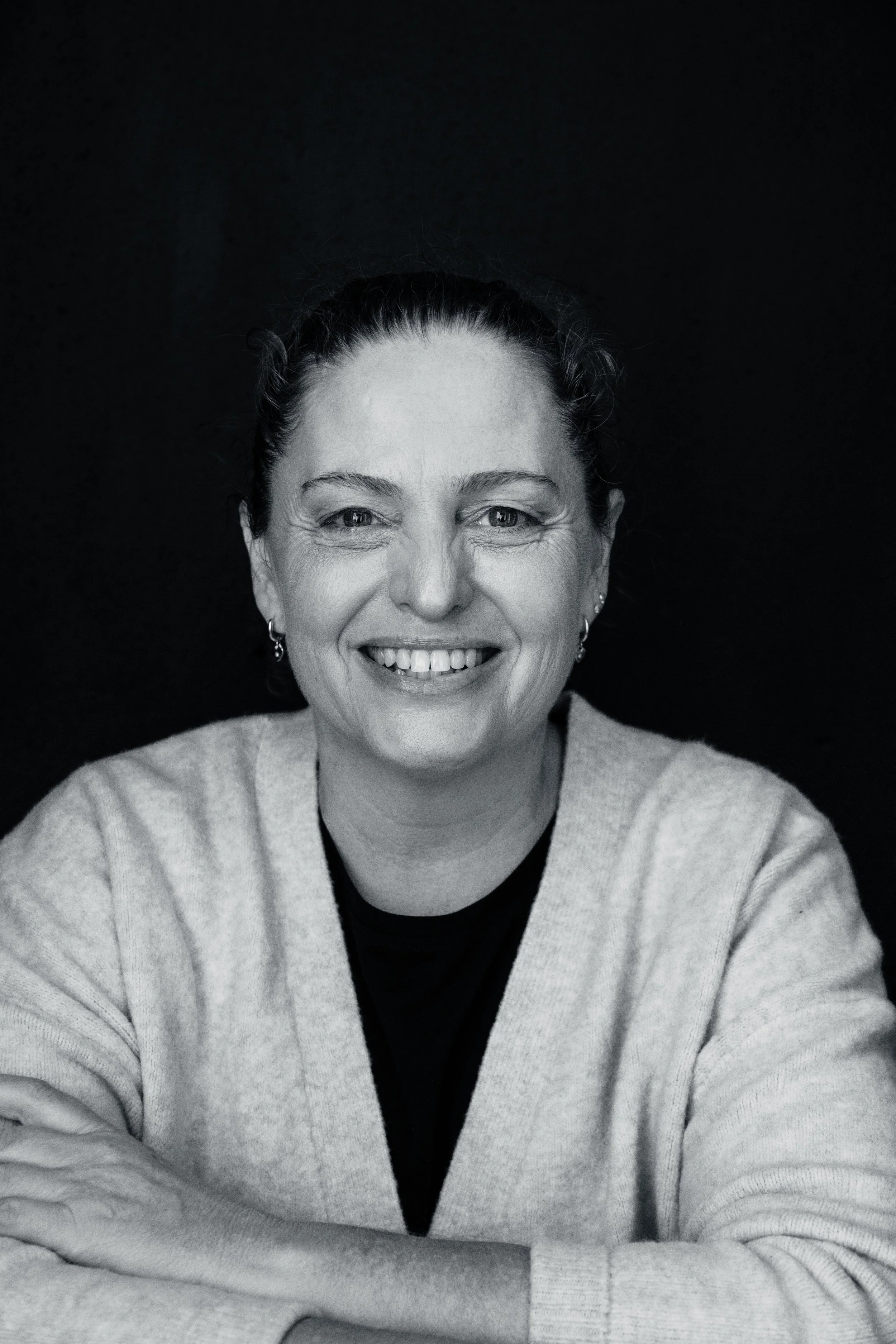 Black and white portrait of a woman smiling, wearing a light-colored sweater and earrings, with arms crossed, against a dark background.