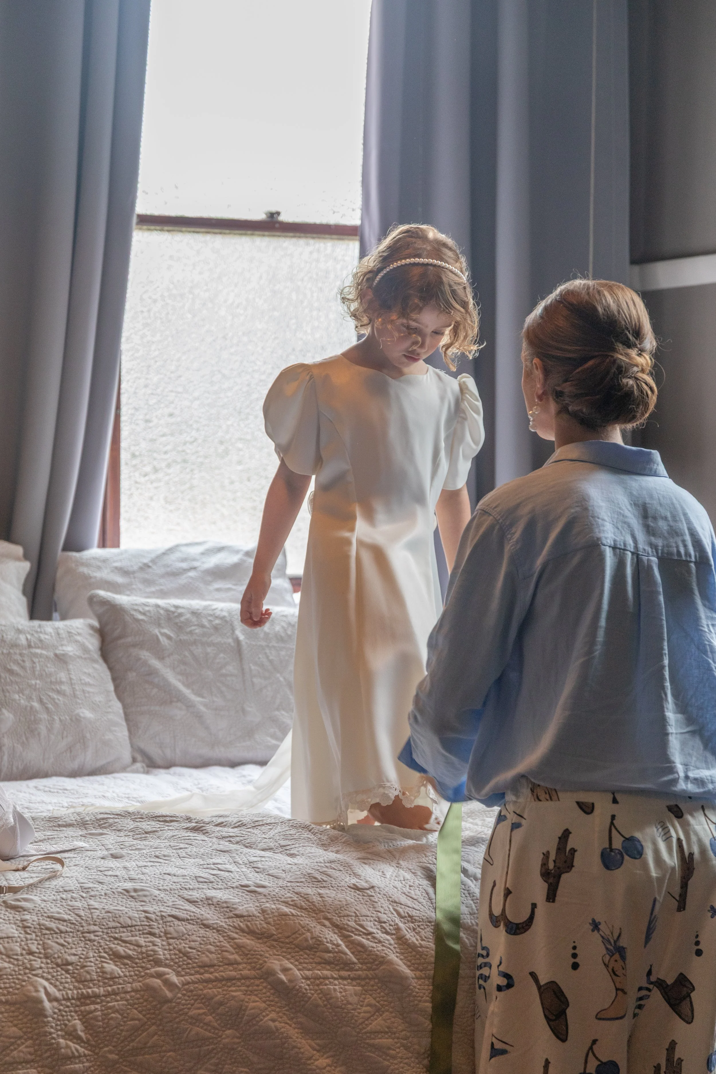 A young girl in a white dress standing on a bed, looking down, while an older woman in pajamas kneels in front of her, holding a ribbon. The scene is in a bedroom with a window and gray curtains.